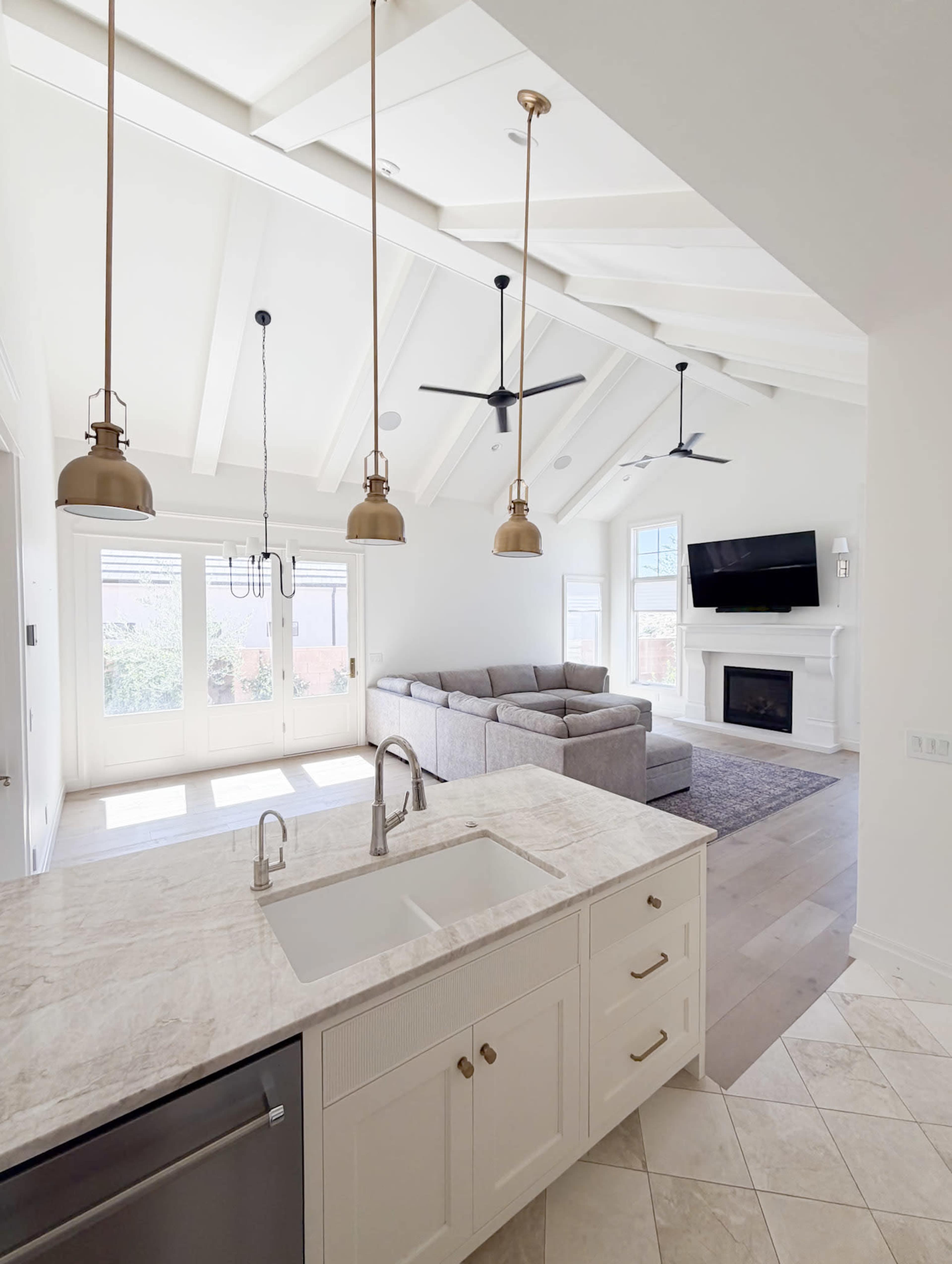 The image shows a modern kitchen with a marble island, pendant lights, and a view into a bright living area featuring a gray couch and a fireplace.