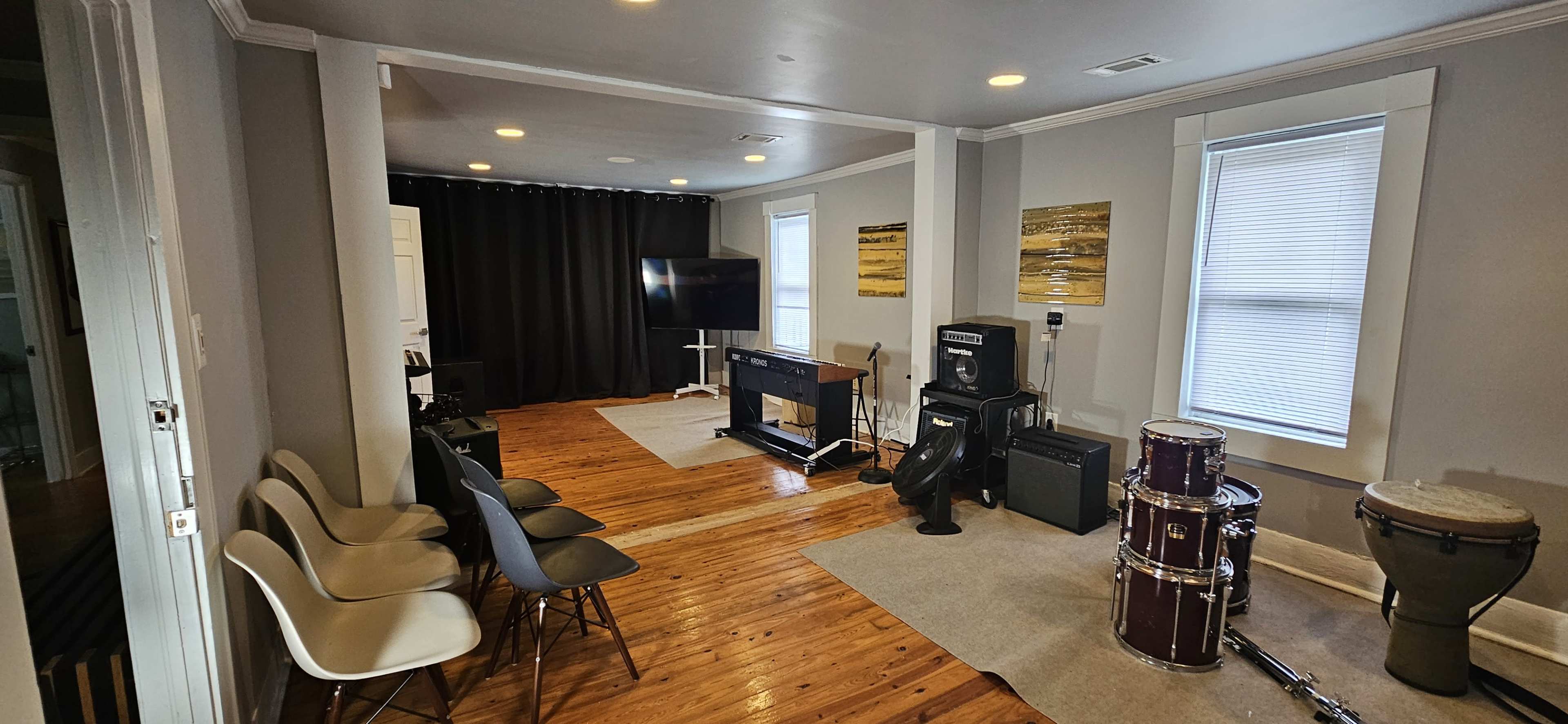 The room features a music rehearsal setup with chairs, a television, various instruments, and sound equipment against neutral-colored walls.
