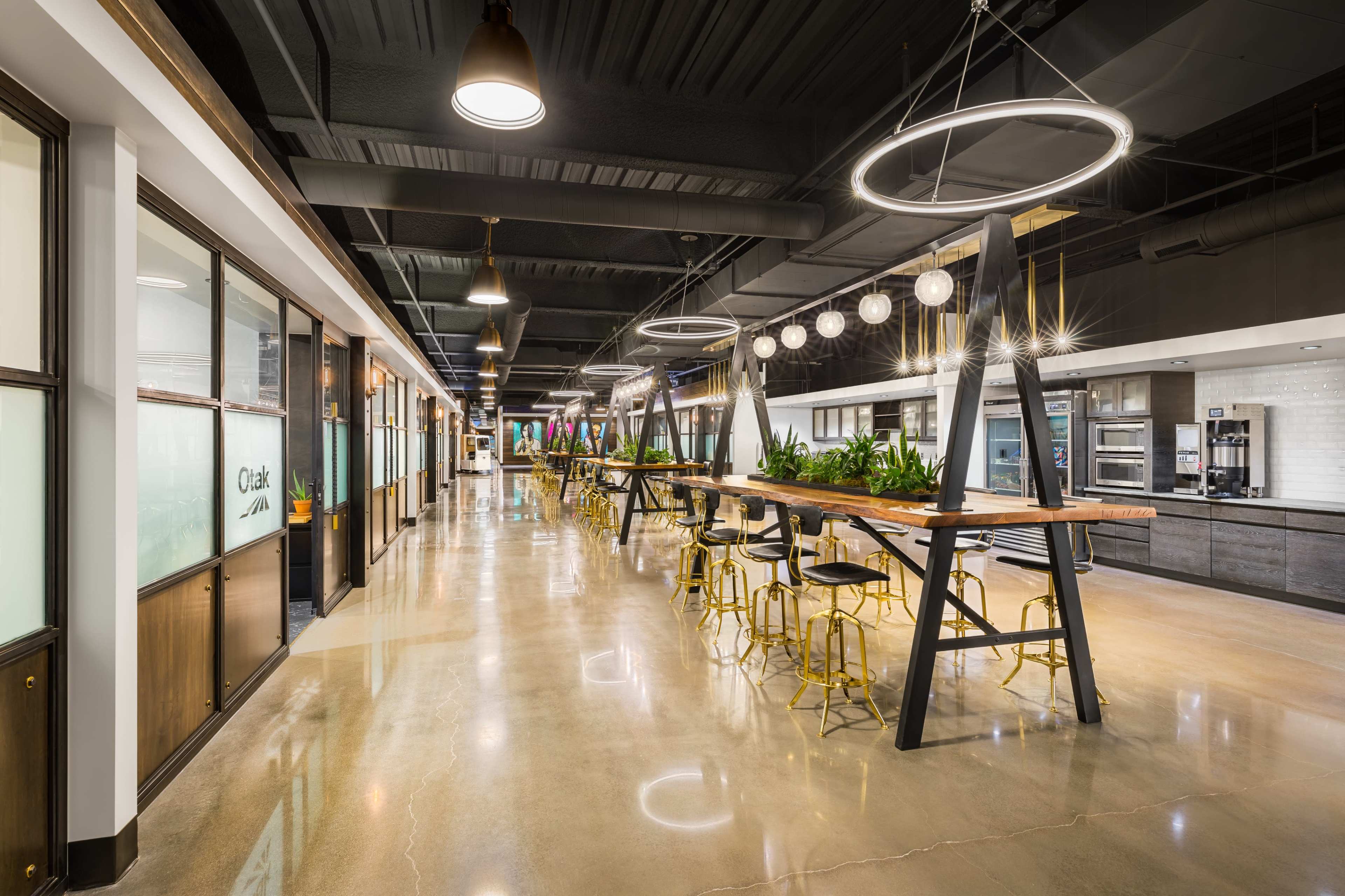 A modern office interior featuring a long communal table, hanging light fixtures, and glass-enclosed meeting rooms along a polished concrete hallway.