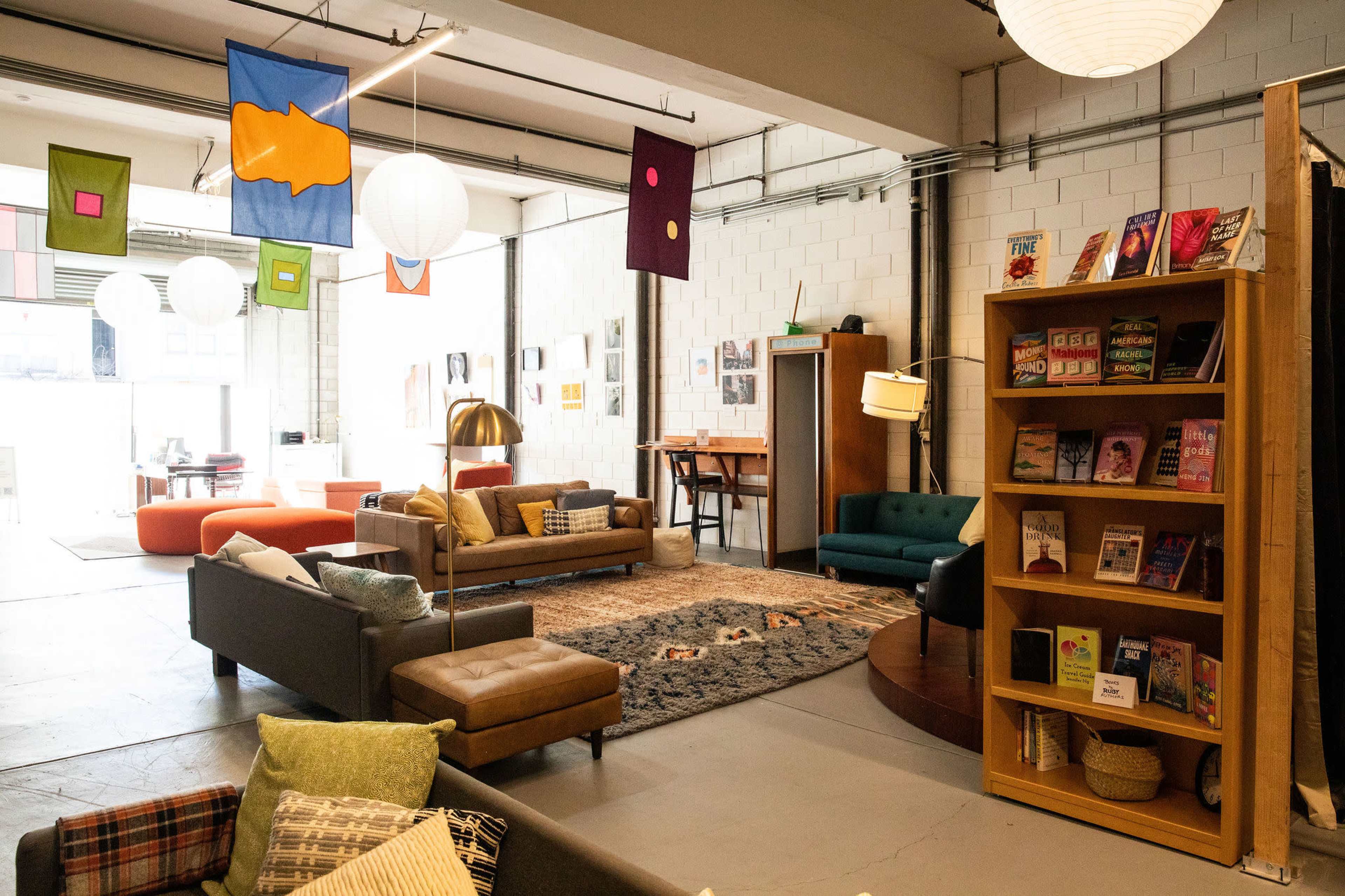 The image shows a spacious, modern lounge area with a mix of colorful furniture, decorative banners hanging from the ceiling, and a bookshelf filled with books.