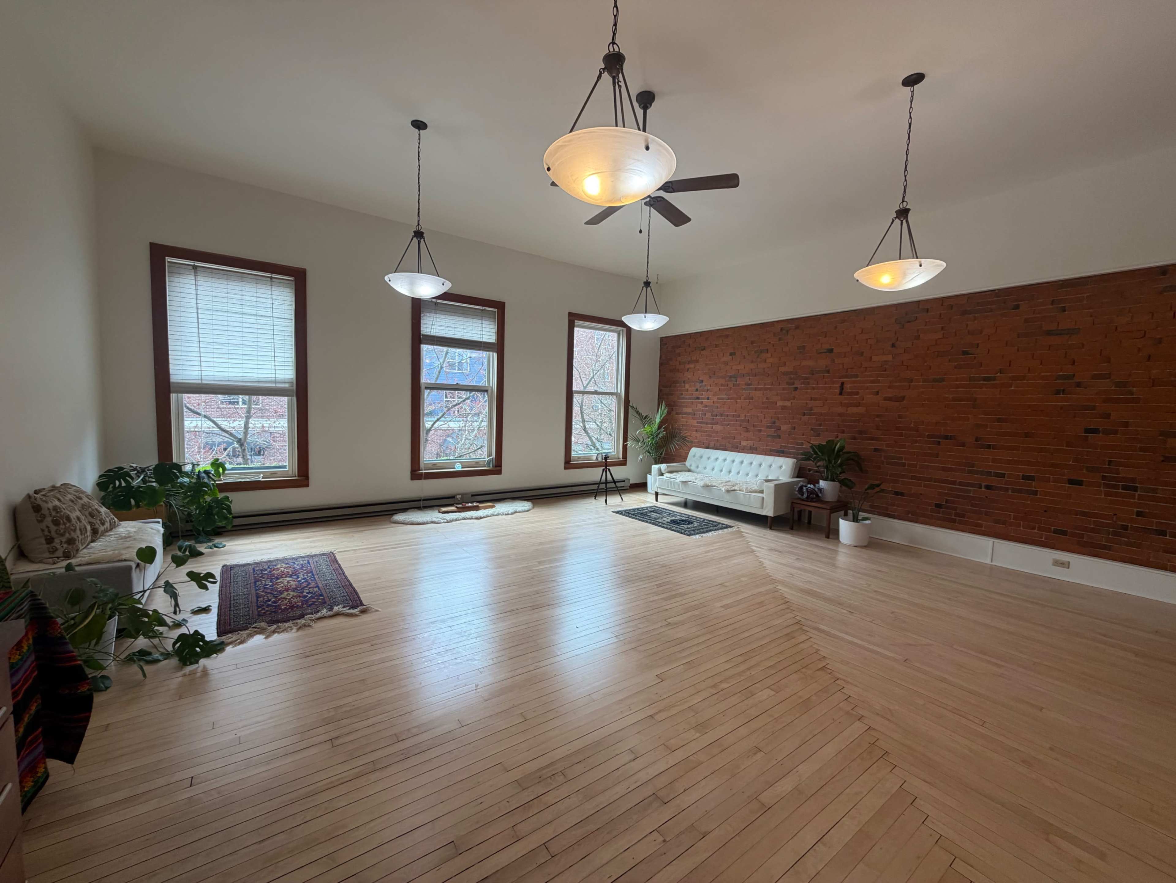 The image shows a spacious room with hardwood floors, three large windows, a brick accent wall, and light fixtures hanging from the ceiling.