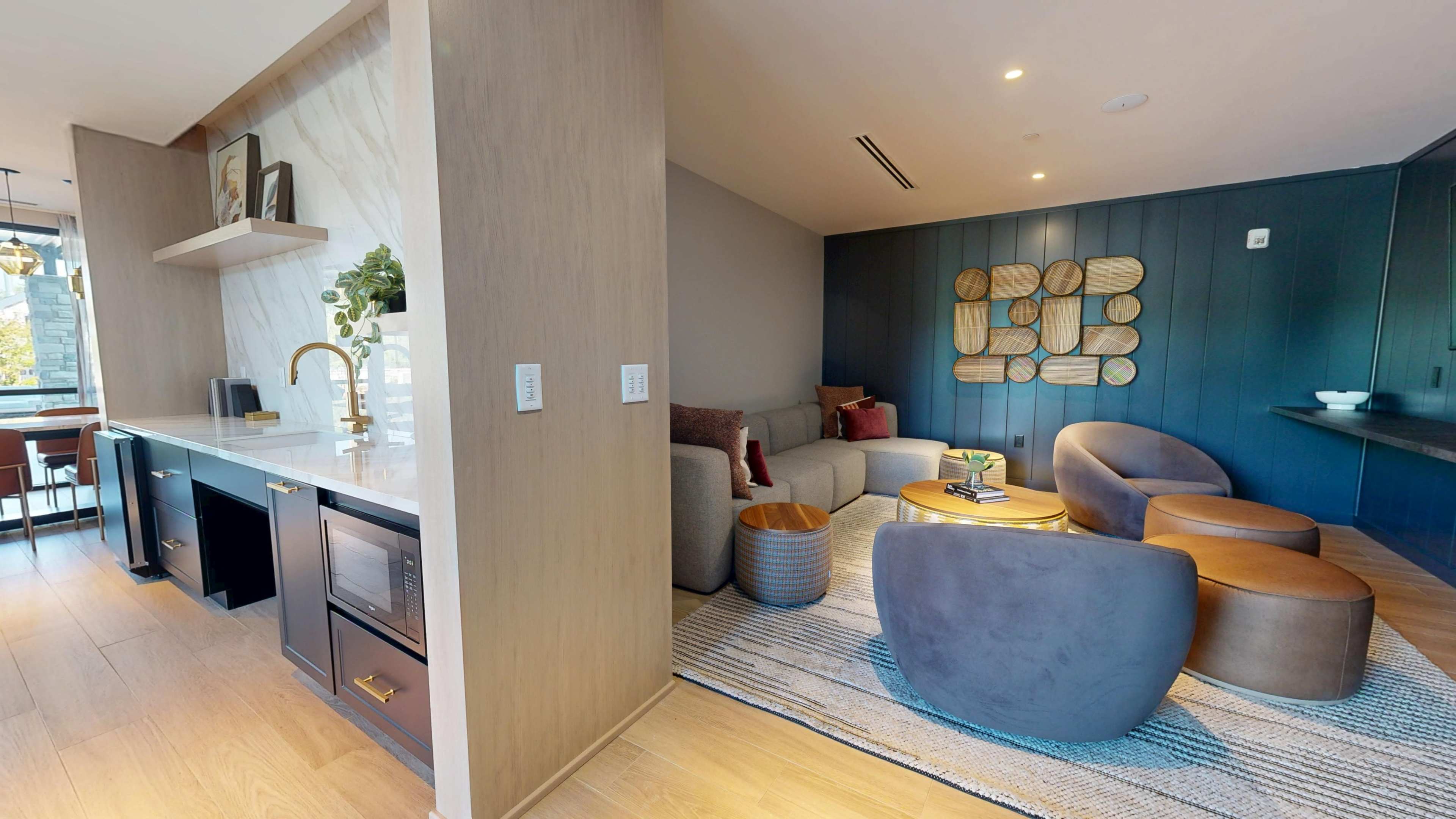 The image shows a modern living space divided into a kitchen area with a countertop and seating, and a cozy lounge area featuring a sofa, accent chairs, and decorative wall art.