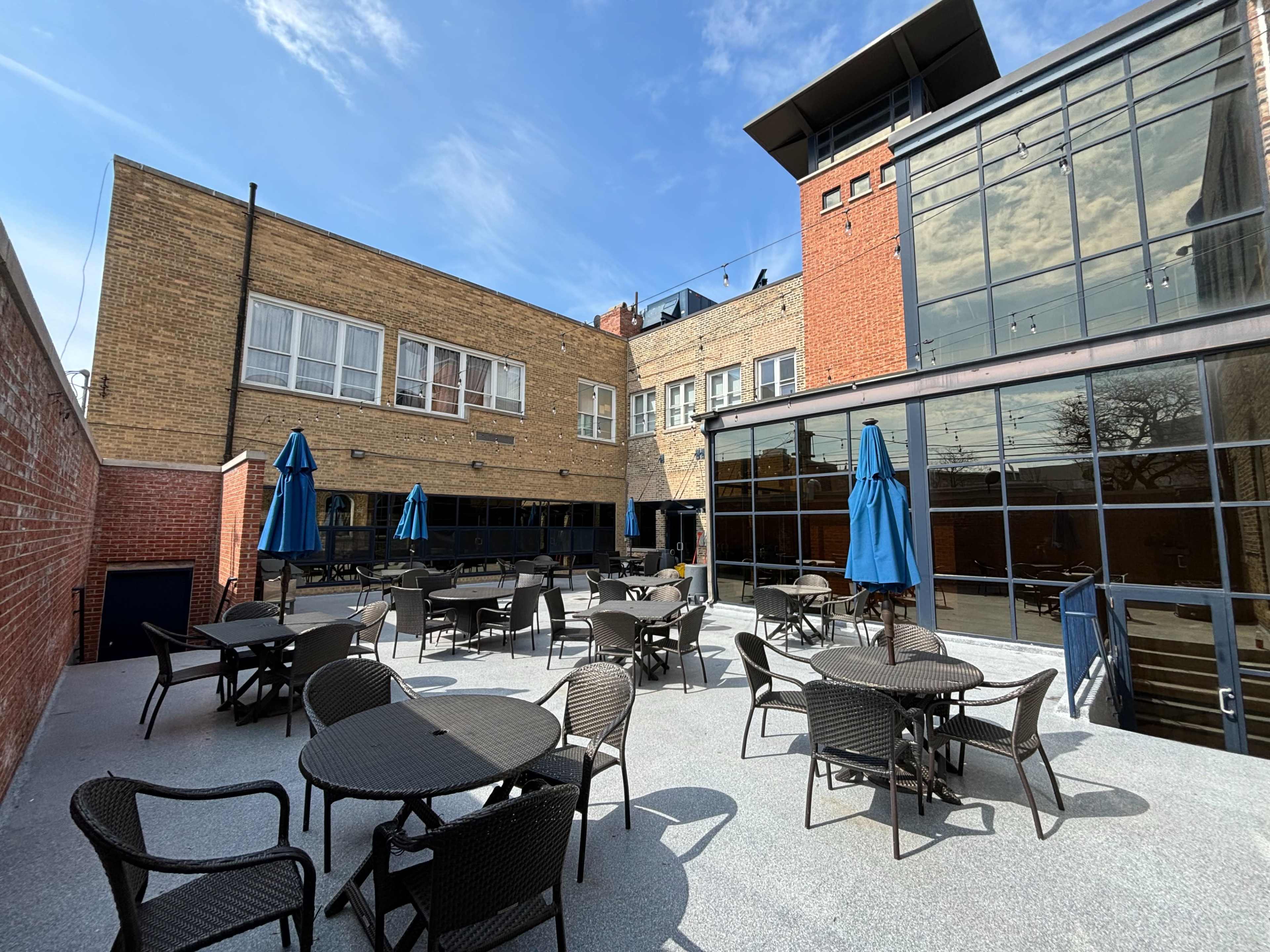 A spacious outdoor patio area with several round tables and blue umbrellas, surrounded by buildings with large glass windows.
