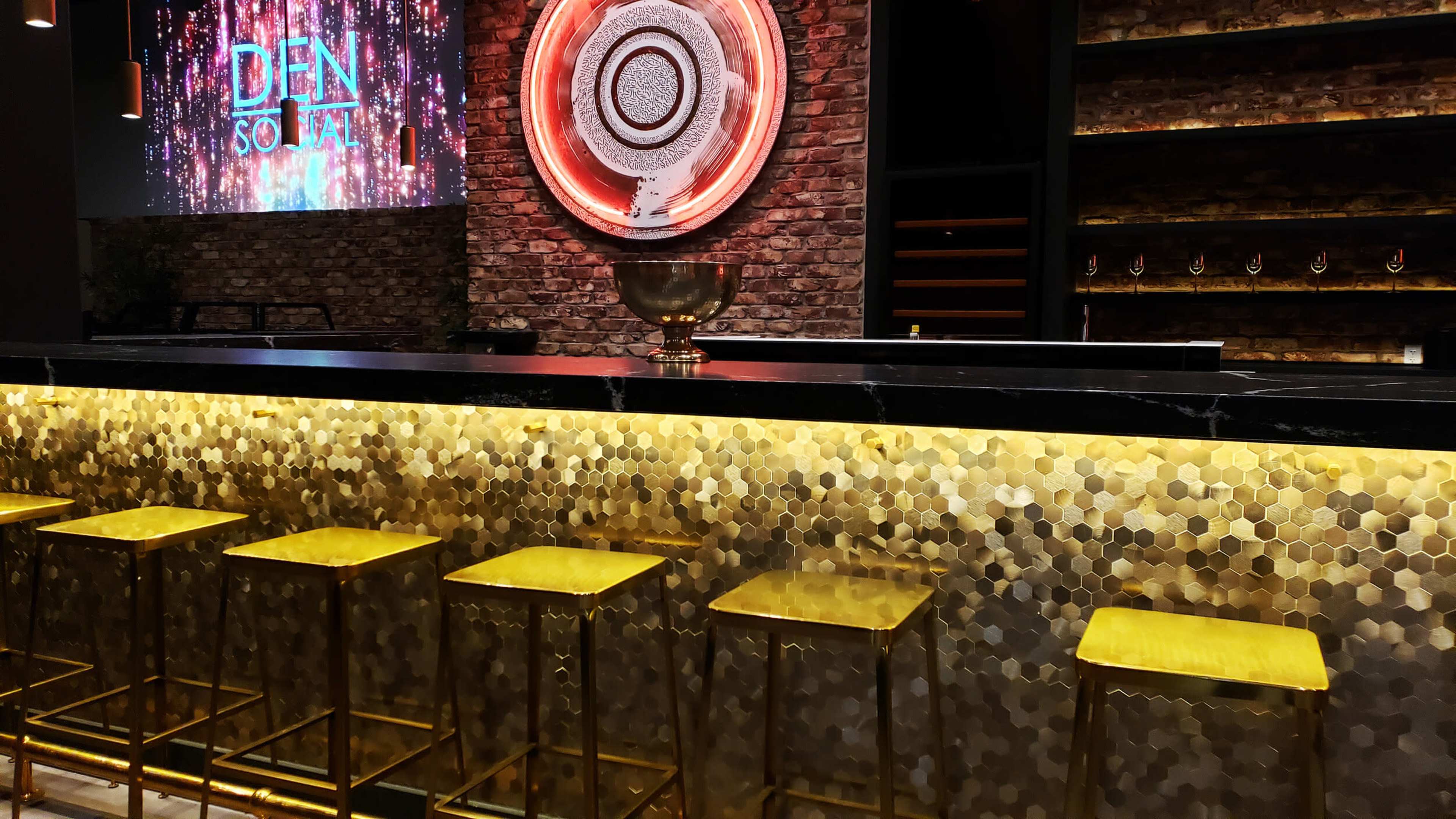 A modern bar with a hexagonal gold tile backsplash, sleek black countertop, and gold stools arranged in front of it, featuring a decorative wall display in the background.