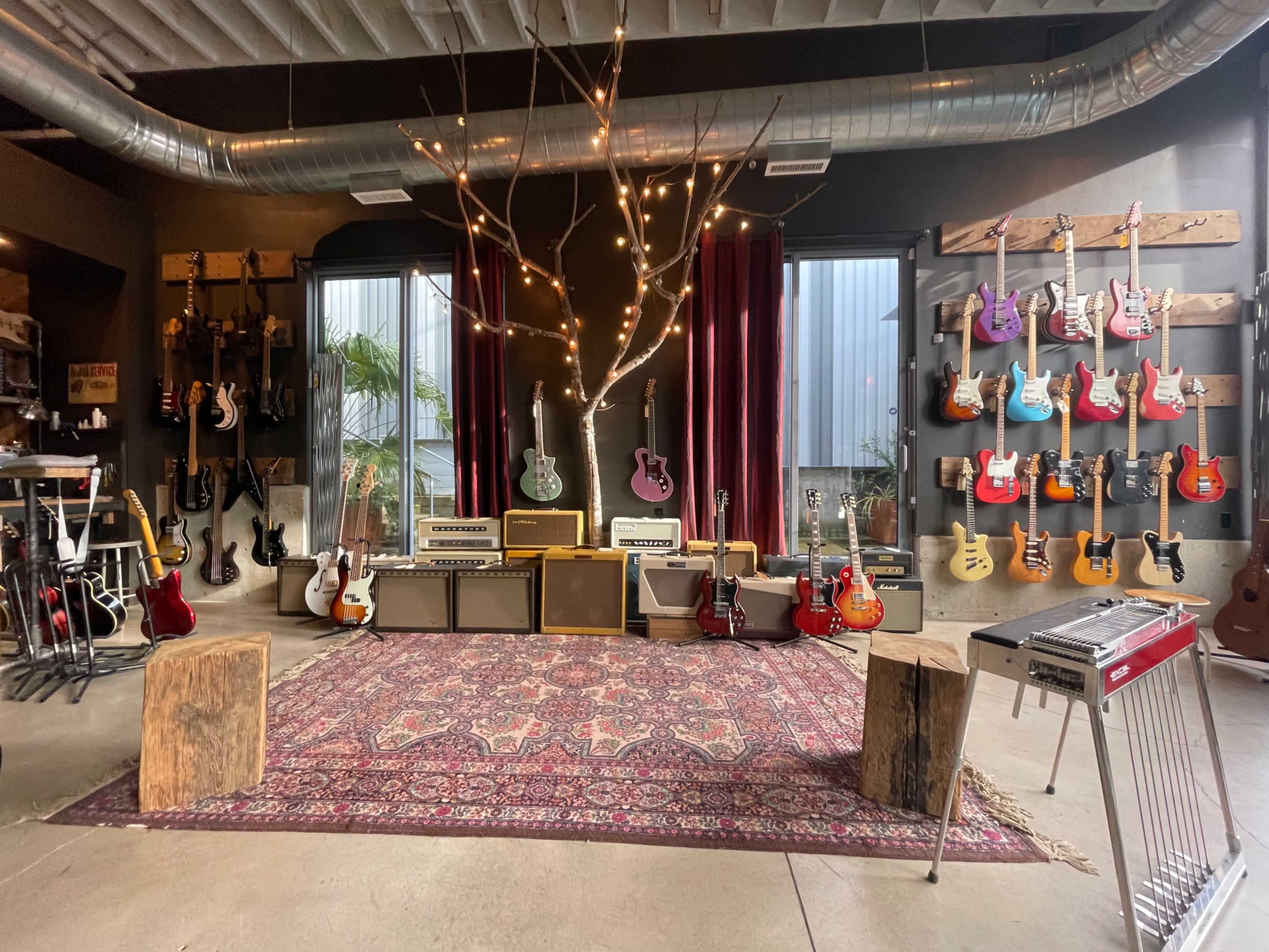 A music studio featuring an array of guitars displayed on walls, amplifiers arranged in a seating area, and a vintage carpet on the floor, complemented by decorative lights and a tree branch.