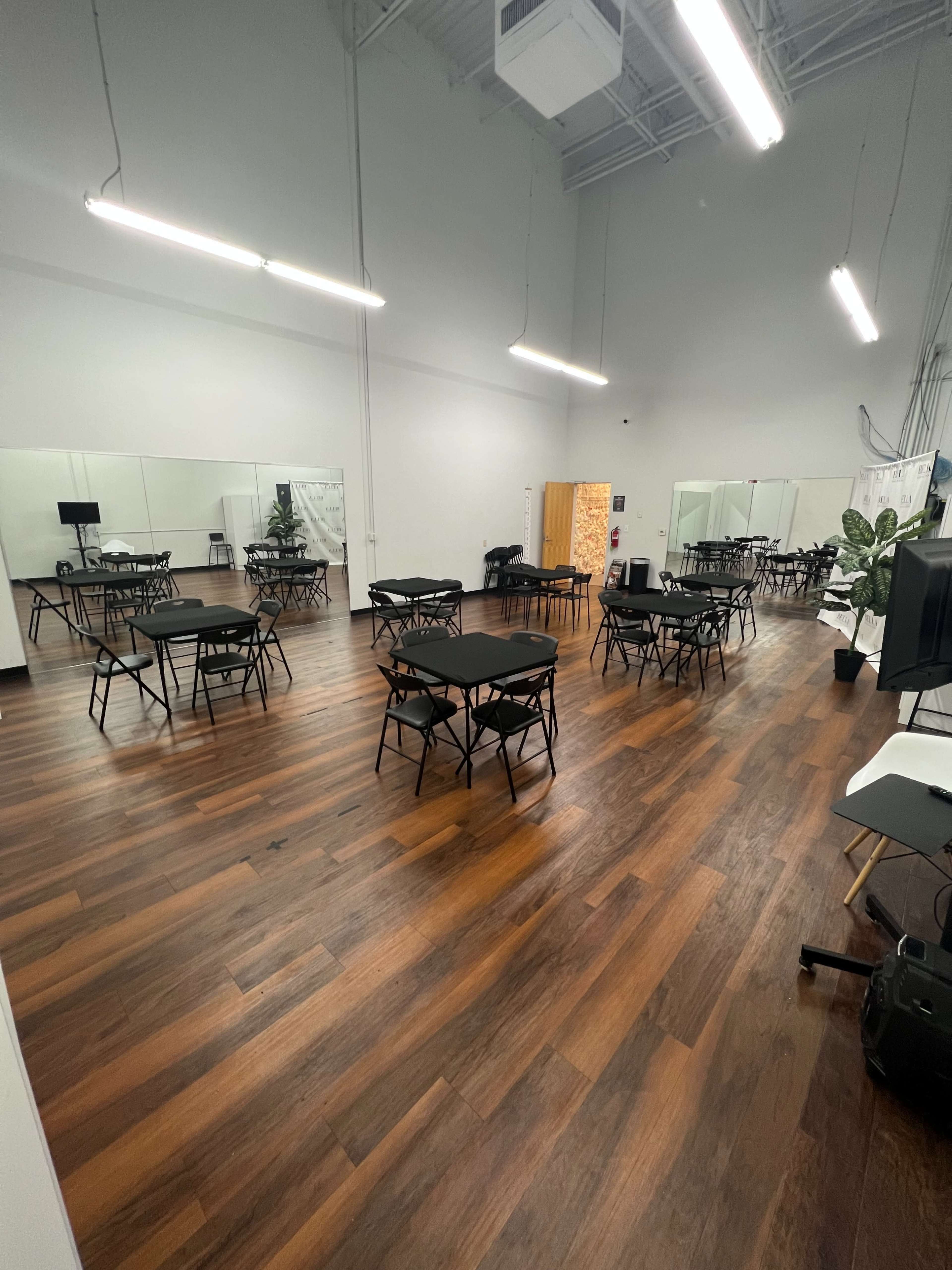 An indoor space with multiple black folding tables arranged in a grid pattern on polished wooden floors, surrounded by mirrors and a few potted plants.