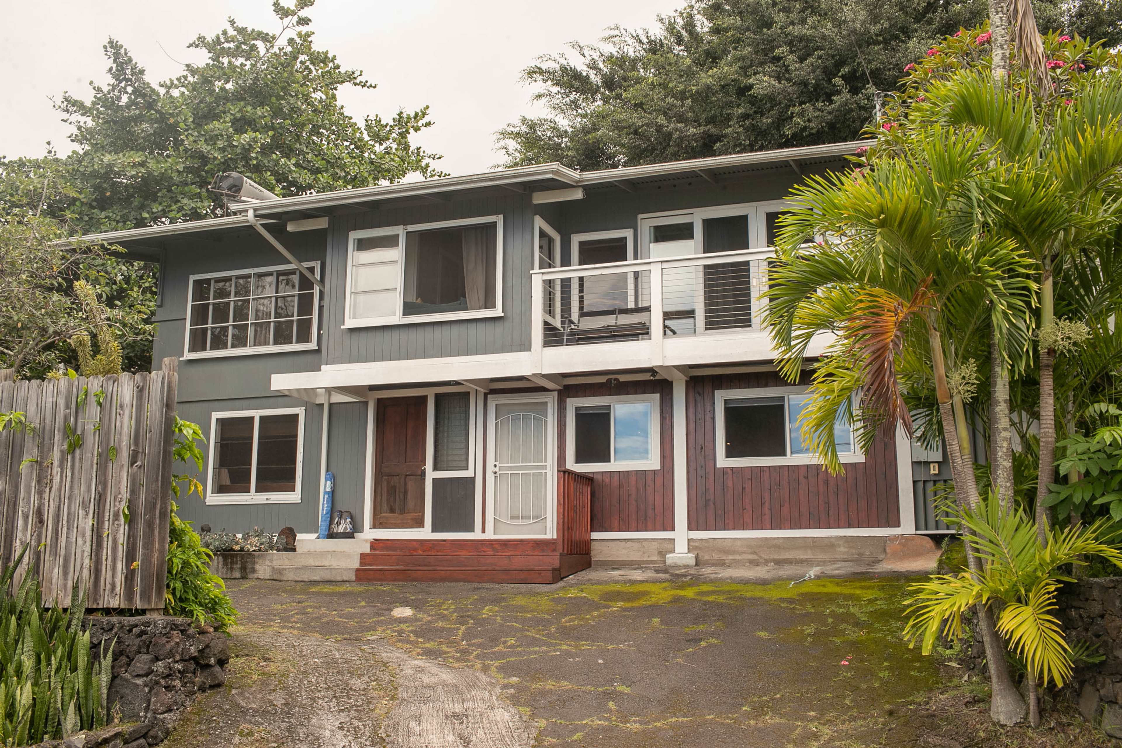 A two-story gray house with a front porch and large windows, surrounded by tropical plants and a gravel driveway.