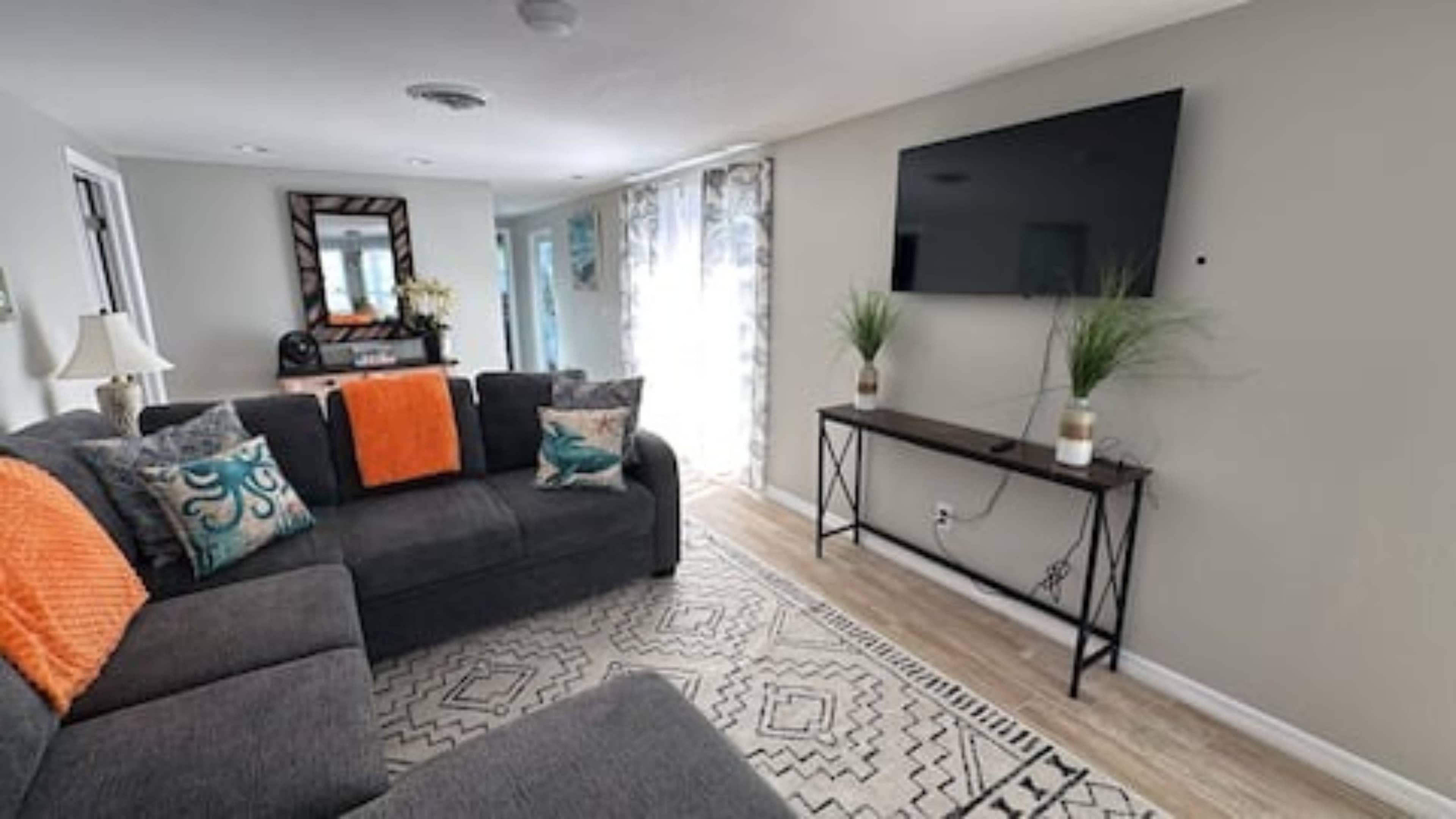 The living room features a gray sectional sofa with colorful cushions, a wall-mounted television, and a decorative console table with potted plants.