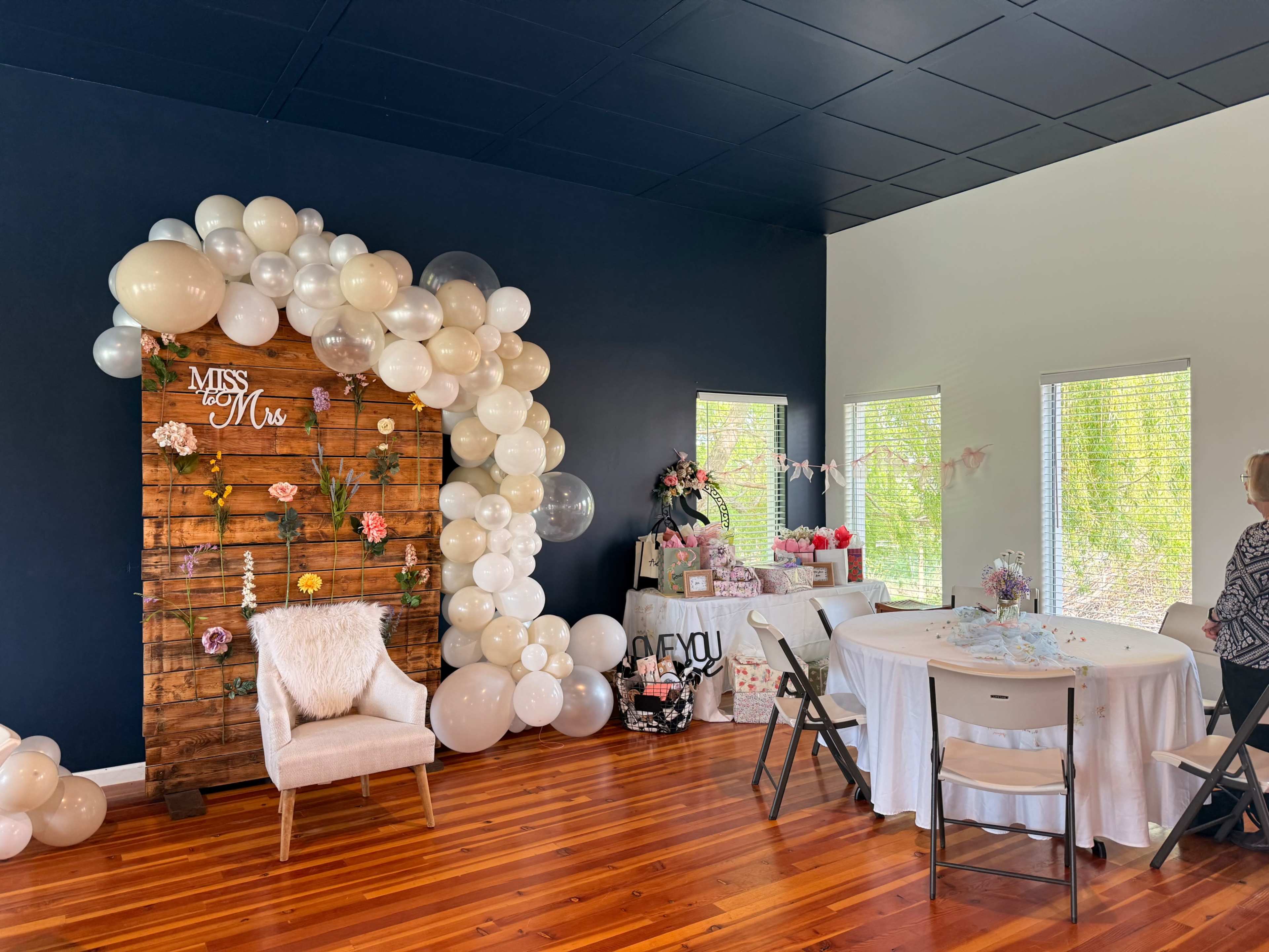 The image shows a decorated indoor space featuring a wooden backdrop with balloons and floral accents, a circular table set for an event, and a woman standing near a display of gifts.