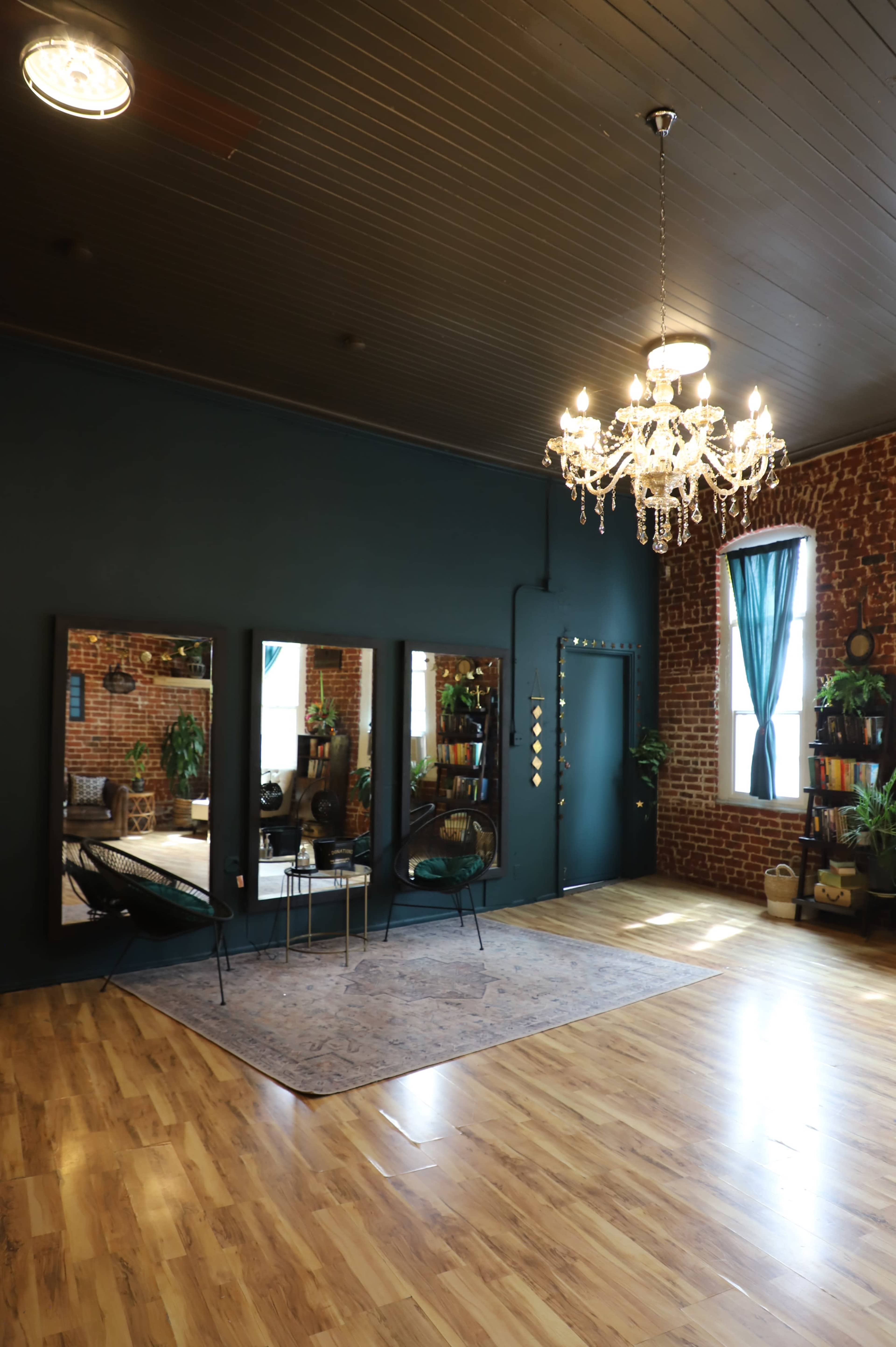 The image shows a spacious room with exposed brick walls, a wooden floor, a large chandelier, three mirrors, and plants arranged around a small seating area.