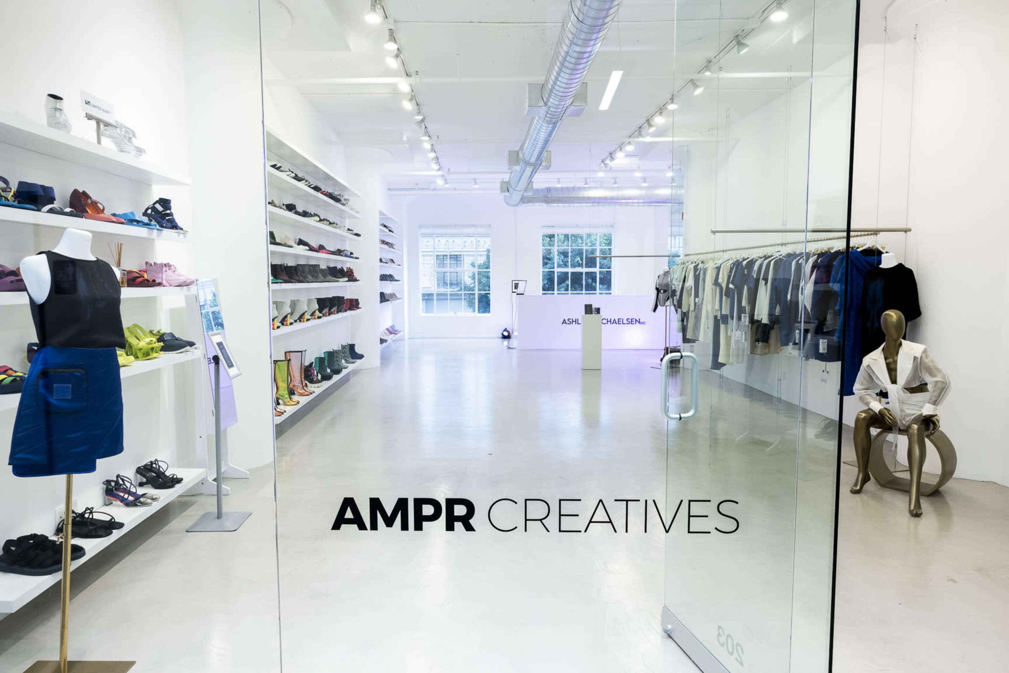 The image shows the entrance to a modern retail space called "AMPR Creatives," featuring a display of clothing and footwear along white shelves and walls.