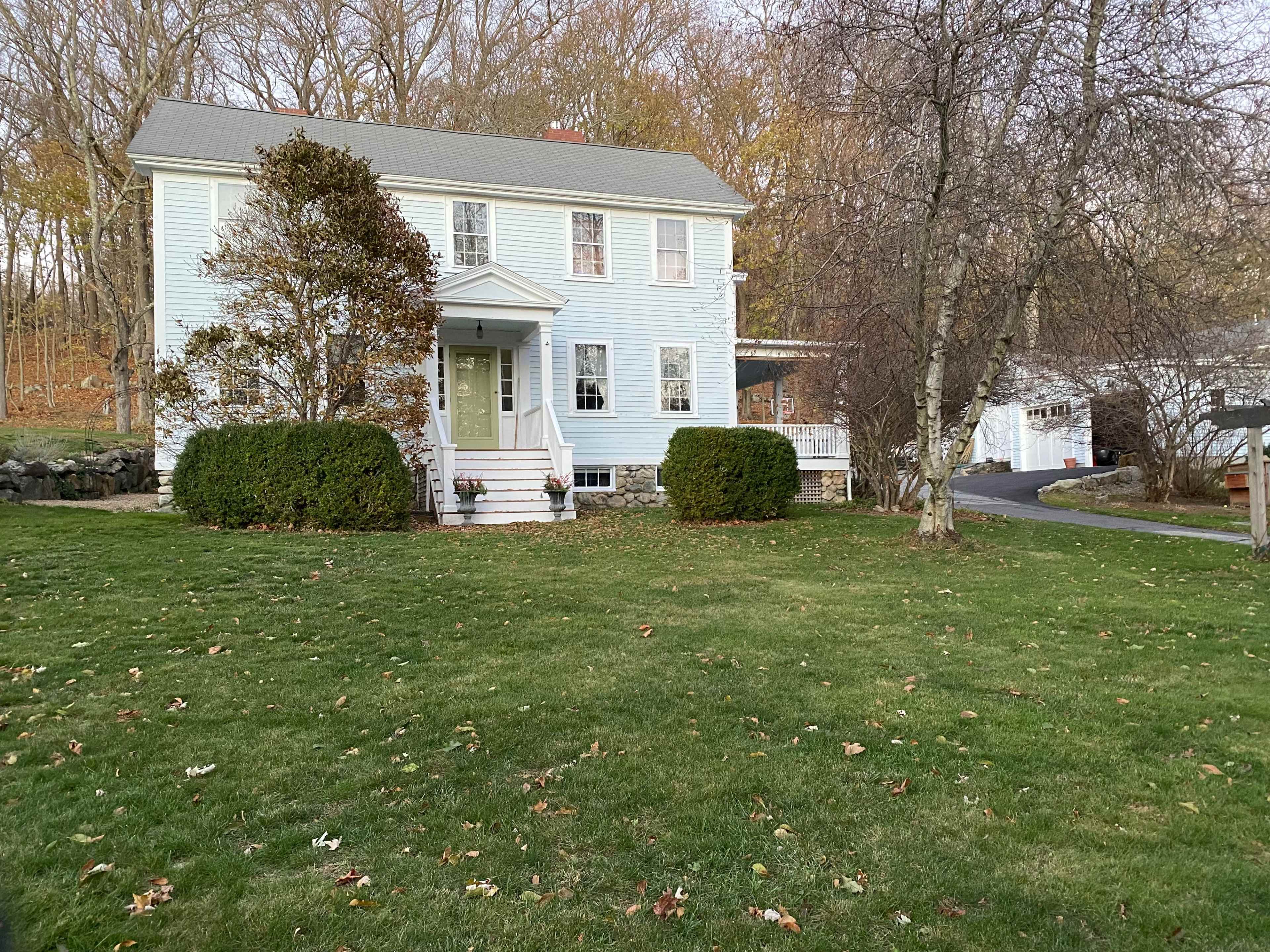 A light blue two-story house with a front porch is set on a grassy lawn surrounded by trees in autumn.