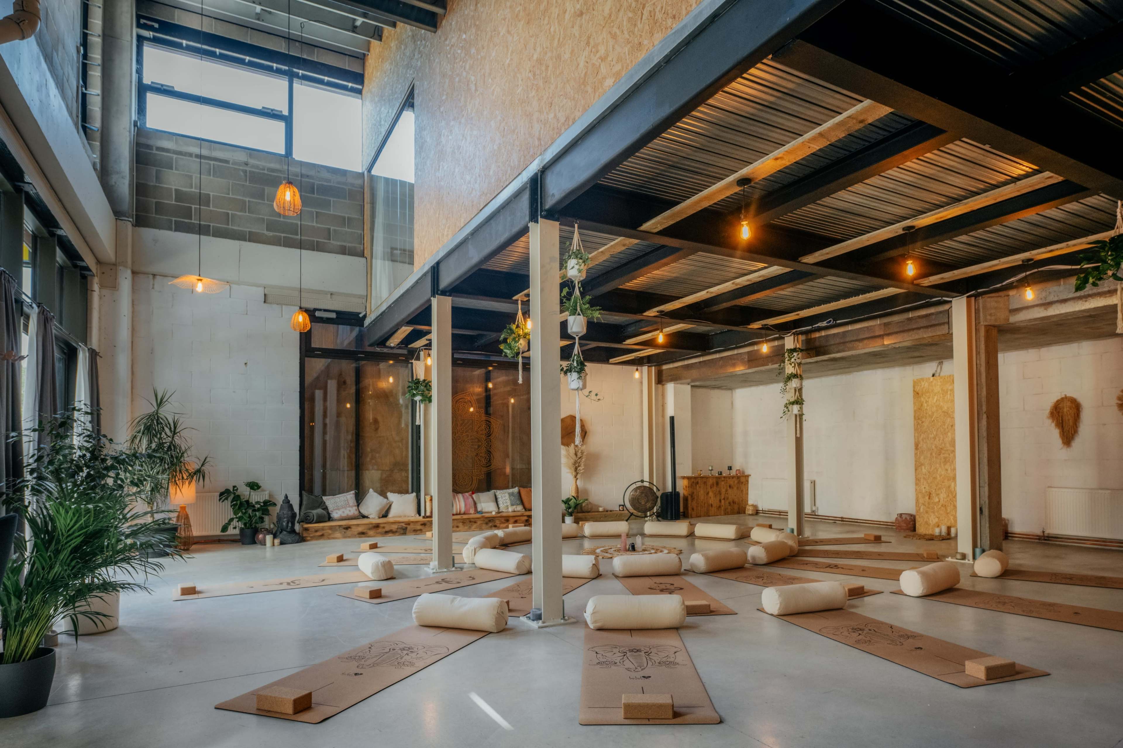 The image shows a spacious yoga studio with wooden beams, yoga mats arranged on the floor, and plants in the corners.