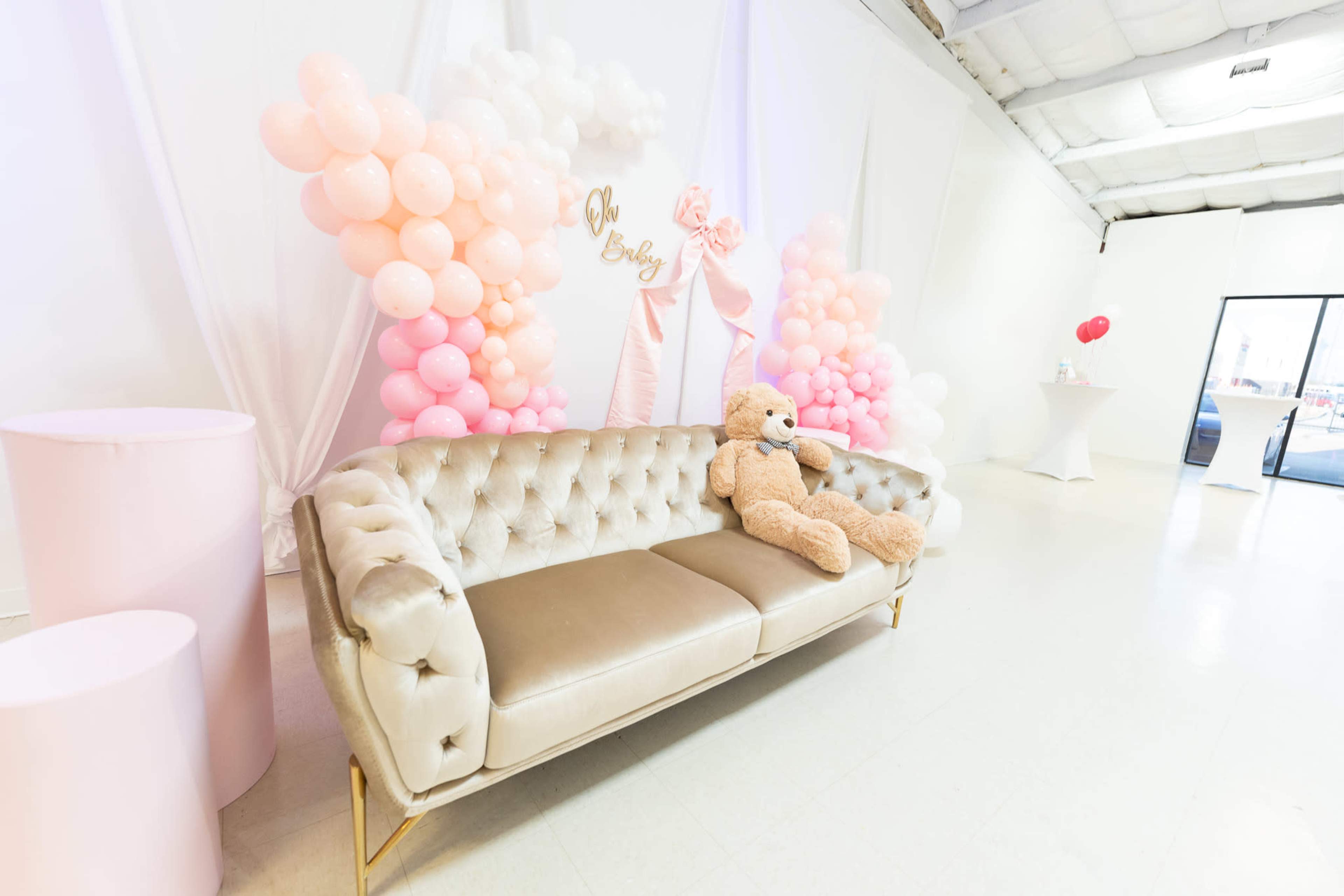 The image shows a plush beige sofa with a teddy bear seated on it, surrounded by decorative pink balloons and a backdrop featuring a bow.