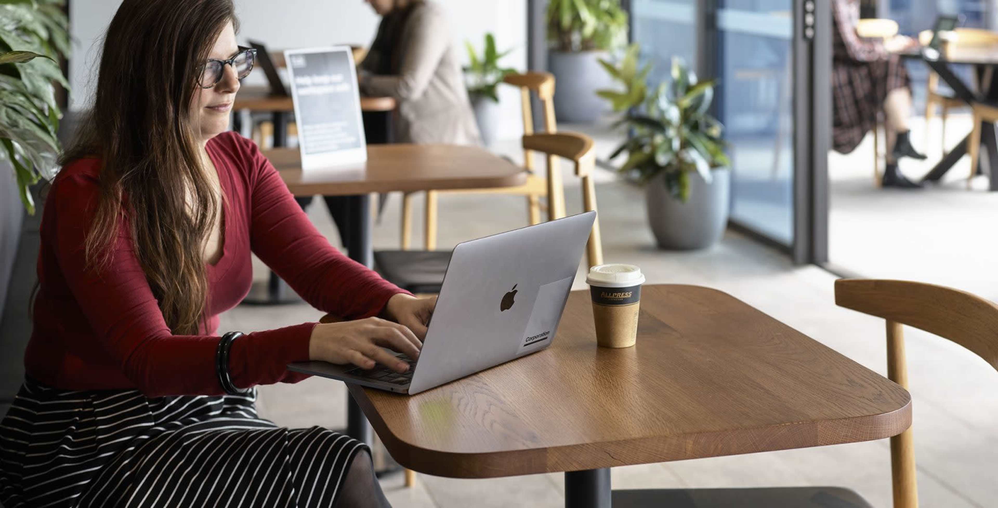 A woman is sitting at a café table working on a laptop while a coffee cup is placed beside her.