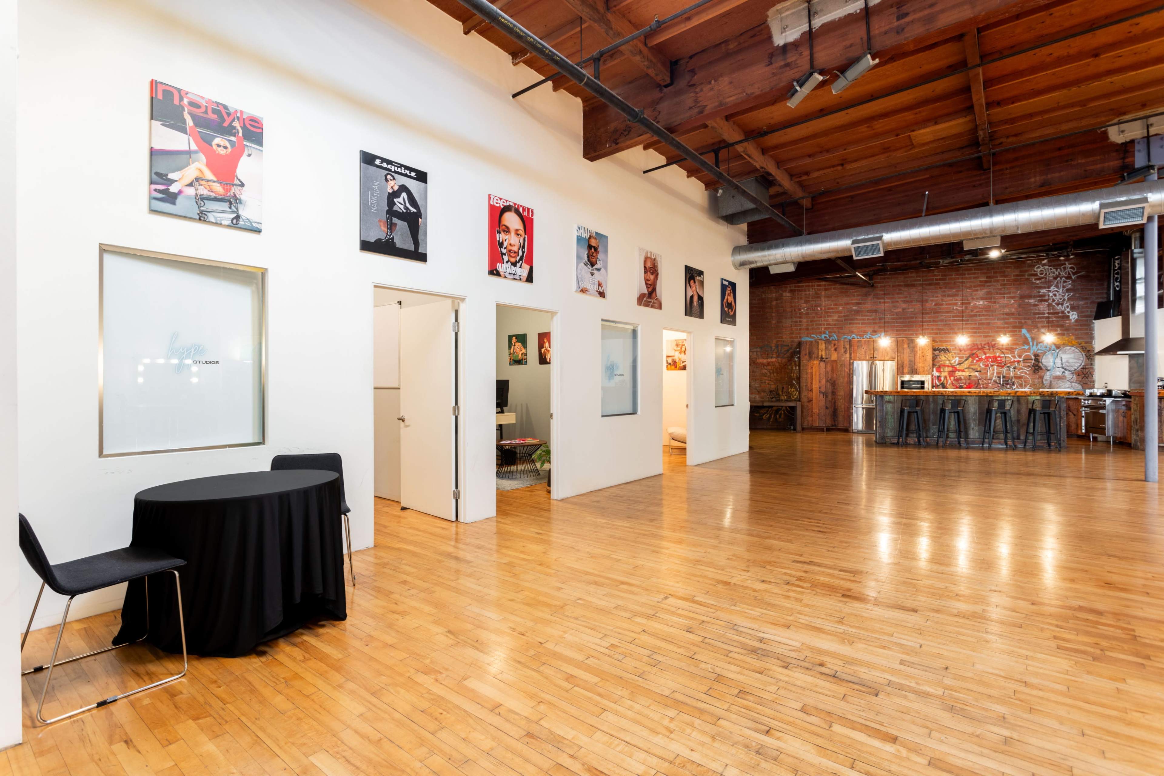 The image shows an open gallery space with wooden floors, white walls displaying framed artwork, and a bar area with high stools in the background.