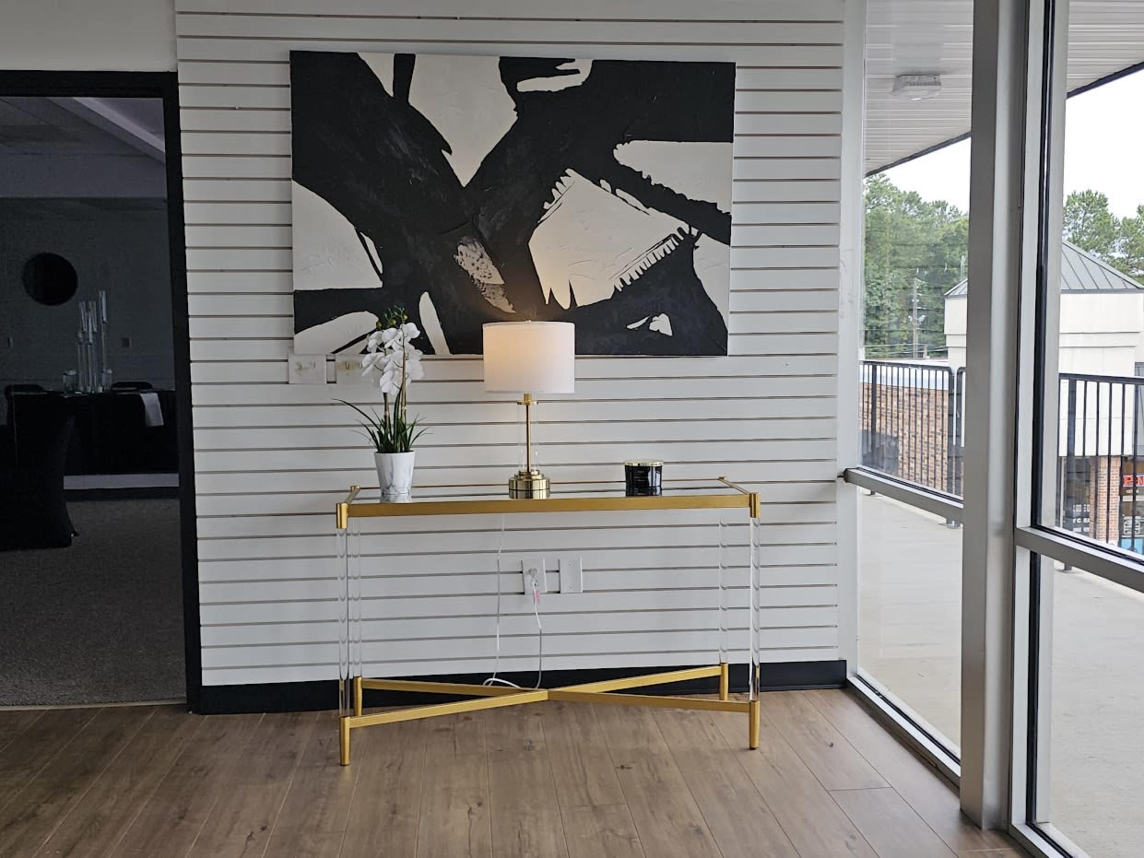 A side table with a lamp and a potted plant, positioned against a wall featuring a large abstract black and white painting.