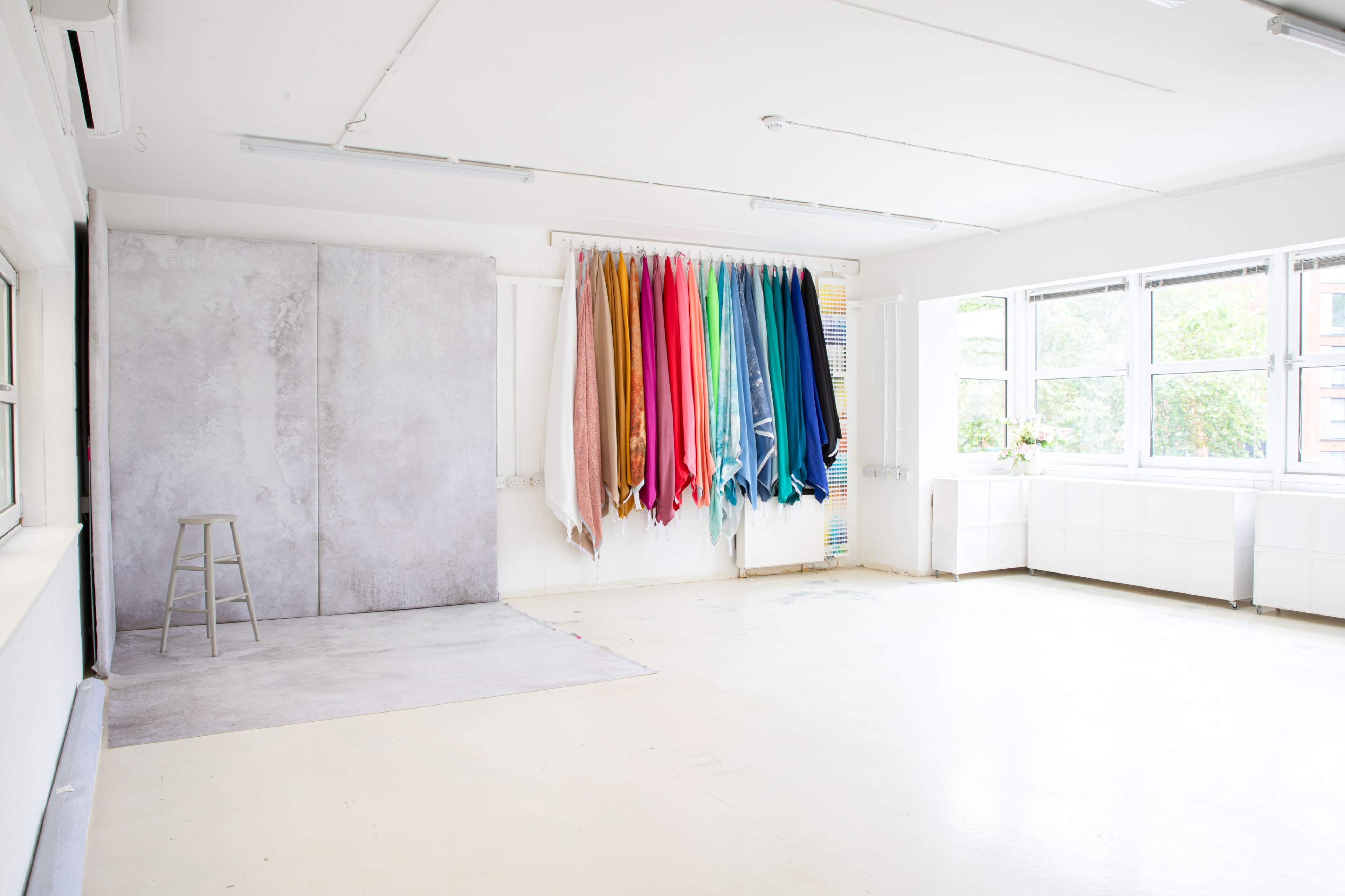 A bright studio space with a gray backdrop and a vertical display of colorful fabric rolls hanging on the wall.