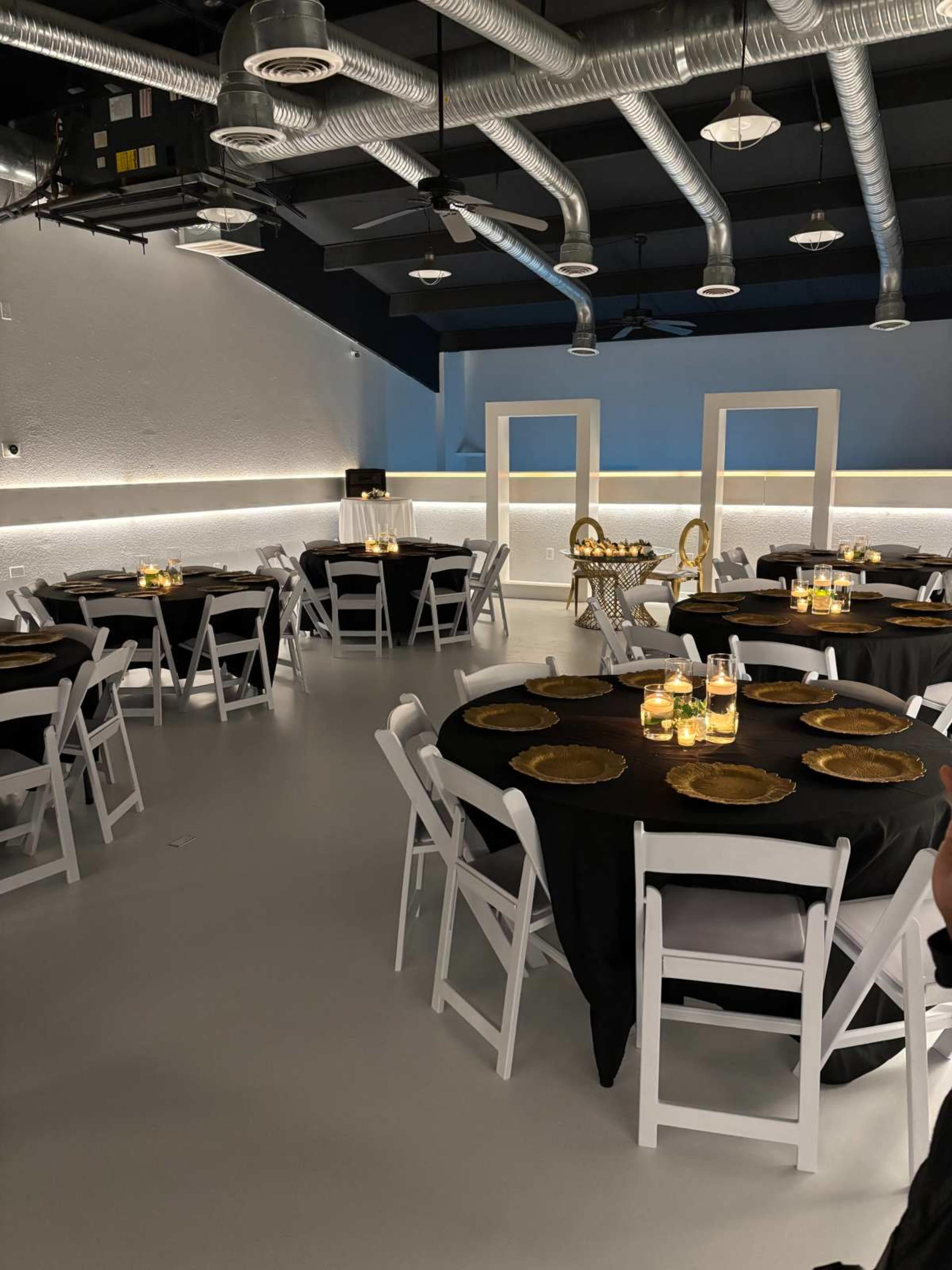 The image shows a modern event space with round tables covered in black tablecloths, each featuring gold chargers and candles, under industrial-style lighting and exposed ductwork.
