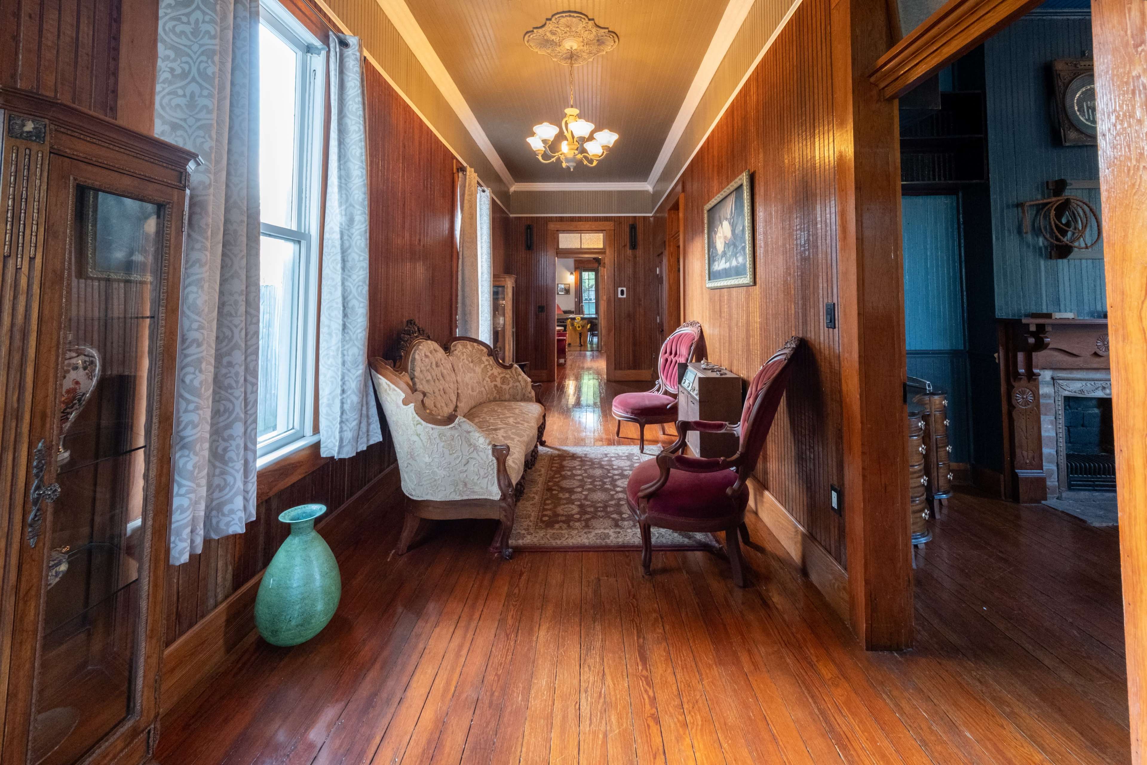 A long hallway features wooden walls, a patterned rug, vintage furniture, and a chandelier.