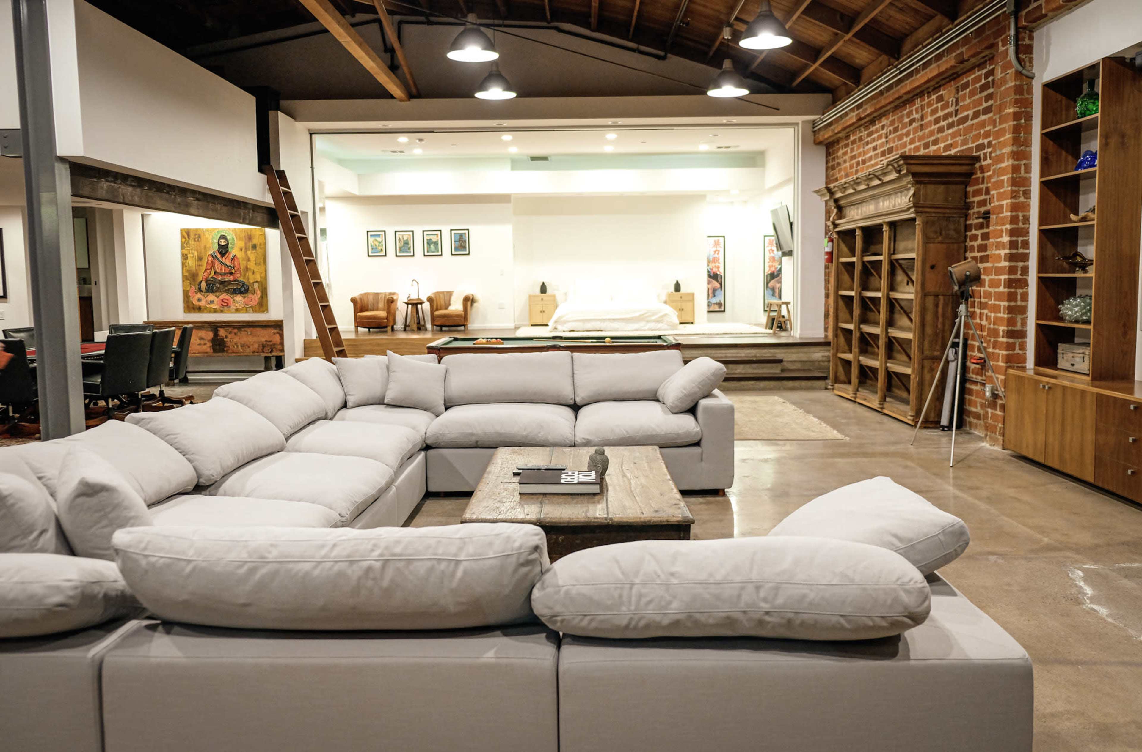 The image shows a spacious, modern living area featuring a large sectional sofa, a coffee table, and an open layout leading to a bedroom area in the background.