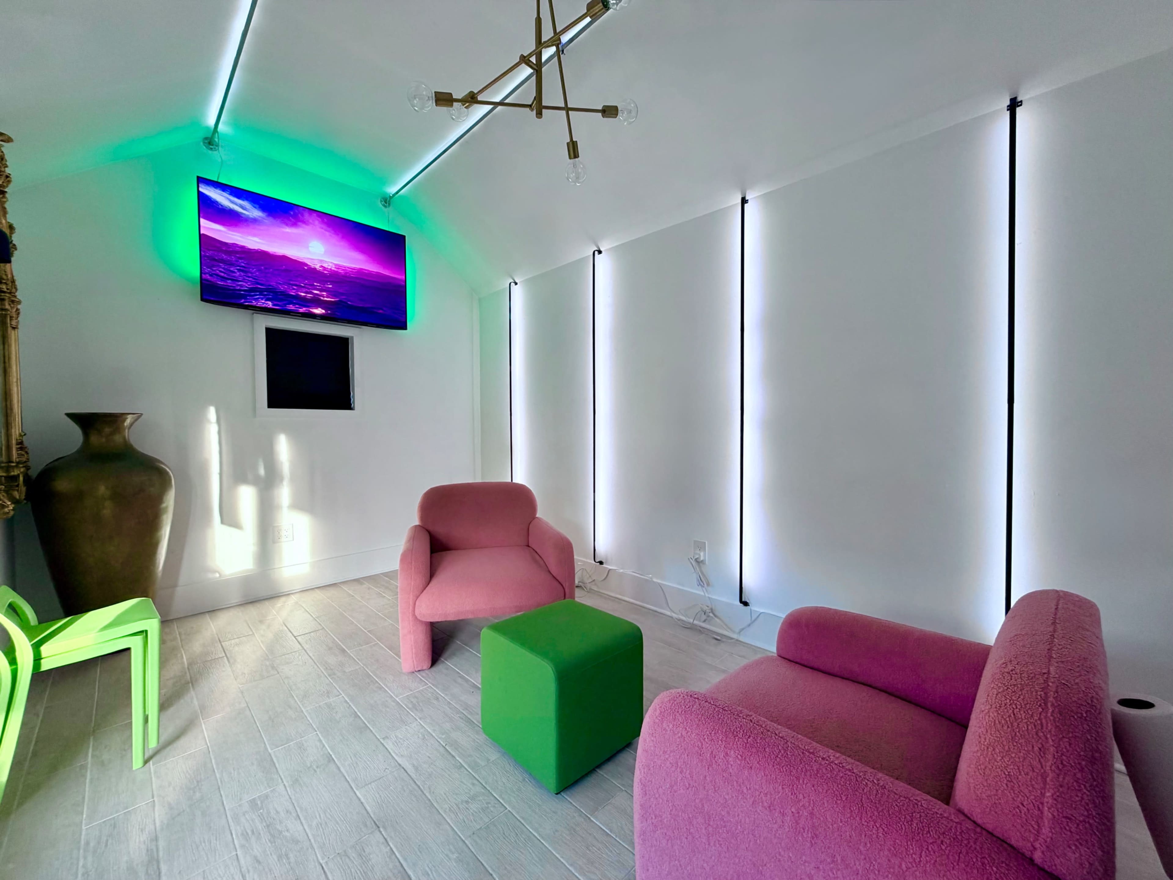 The image shows a modern room with two pink chairs, a green stool, an abstract TV display, and illuminated walls.