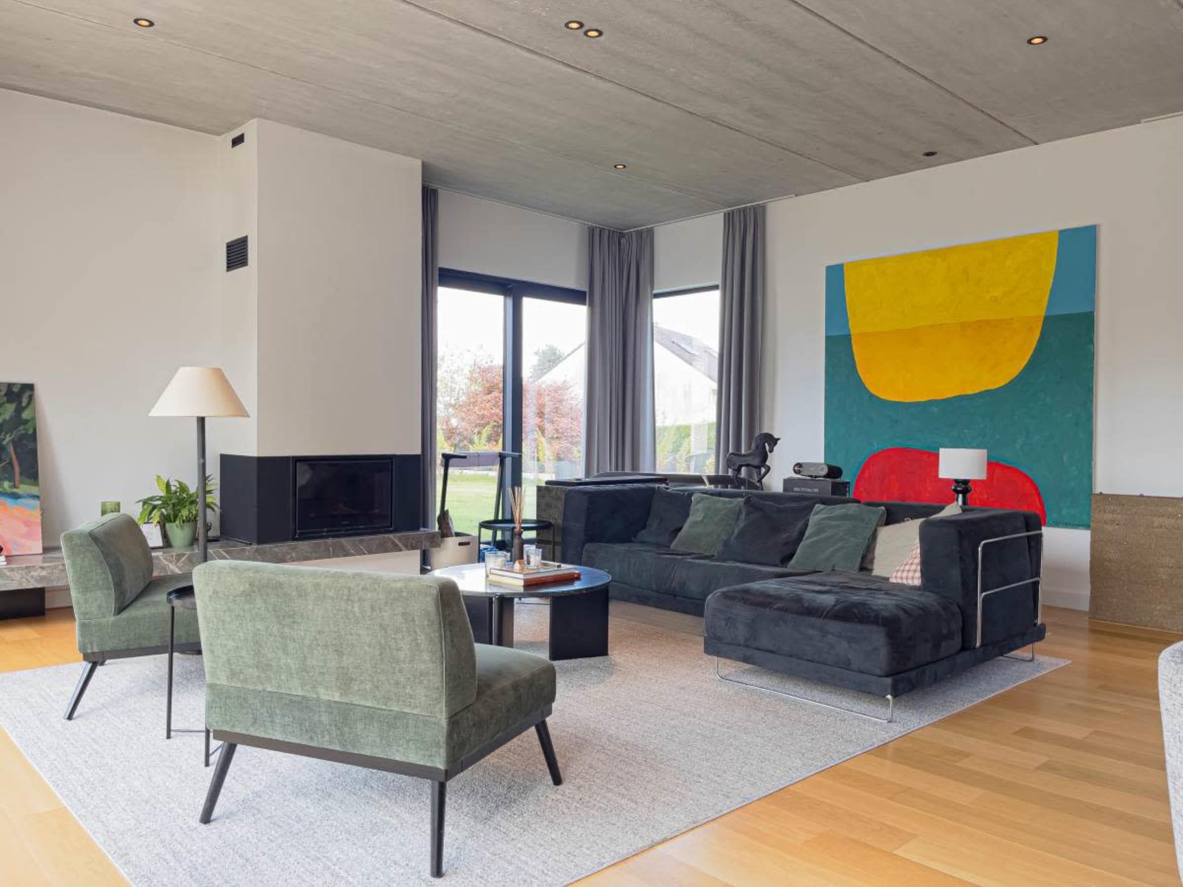 The image depicts a modern living room featuring a large sofa, two armchairs, a coffee table, and a wall art piece, all set against a backdrop of large windows and a concrete ceiling.