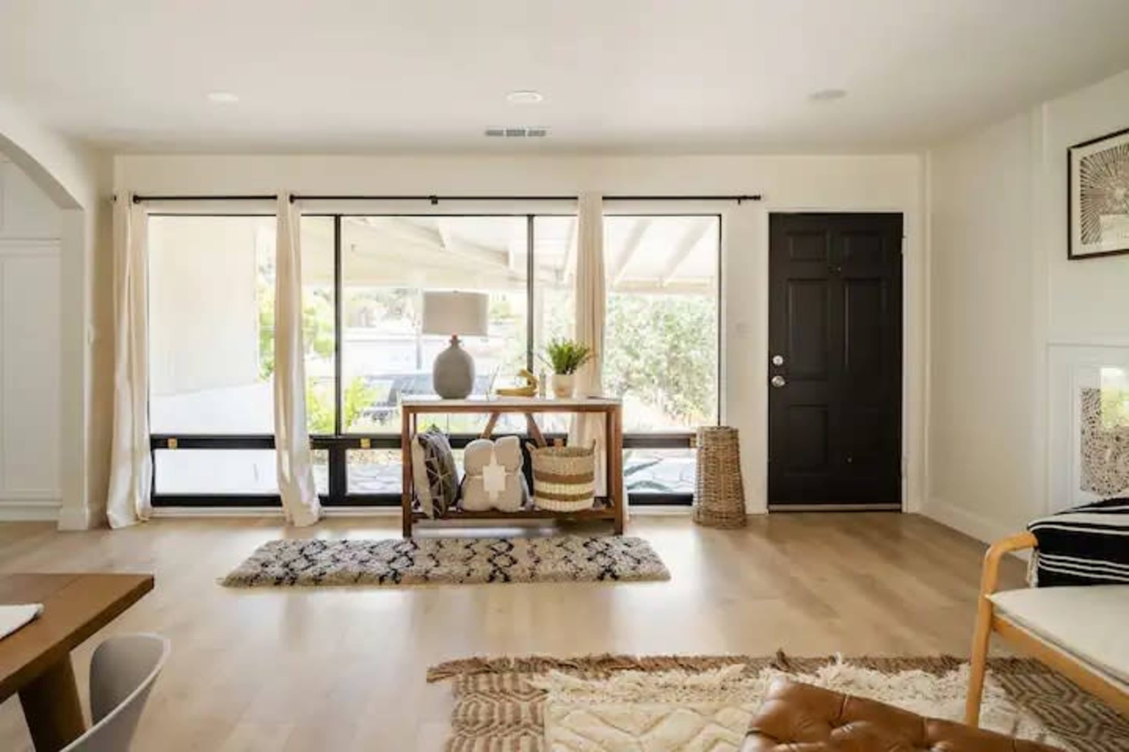 A modern living room with a table displaying a lamp and decorative items, large windows allowing natural light, and sliding doors leading to an outdoor area.