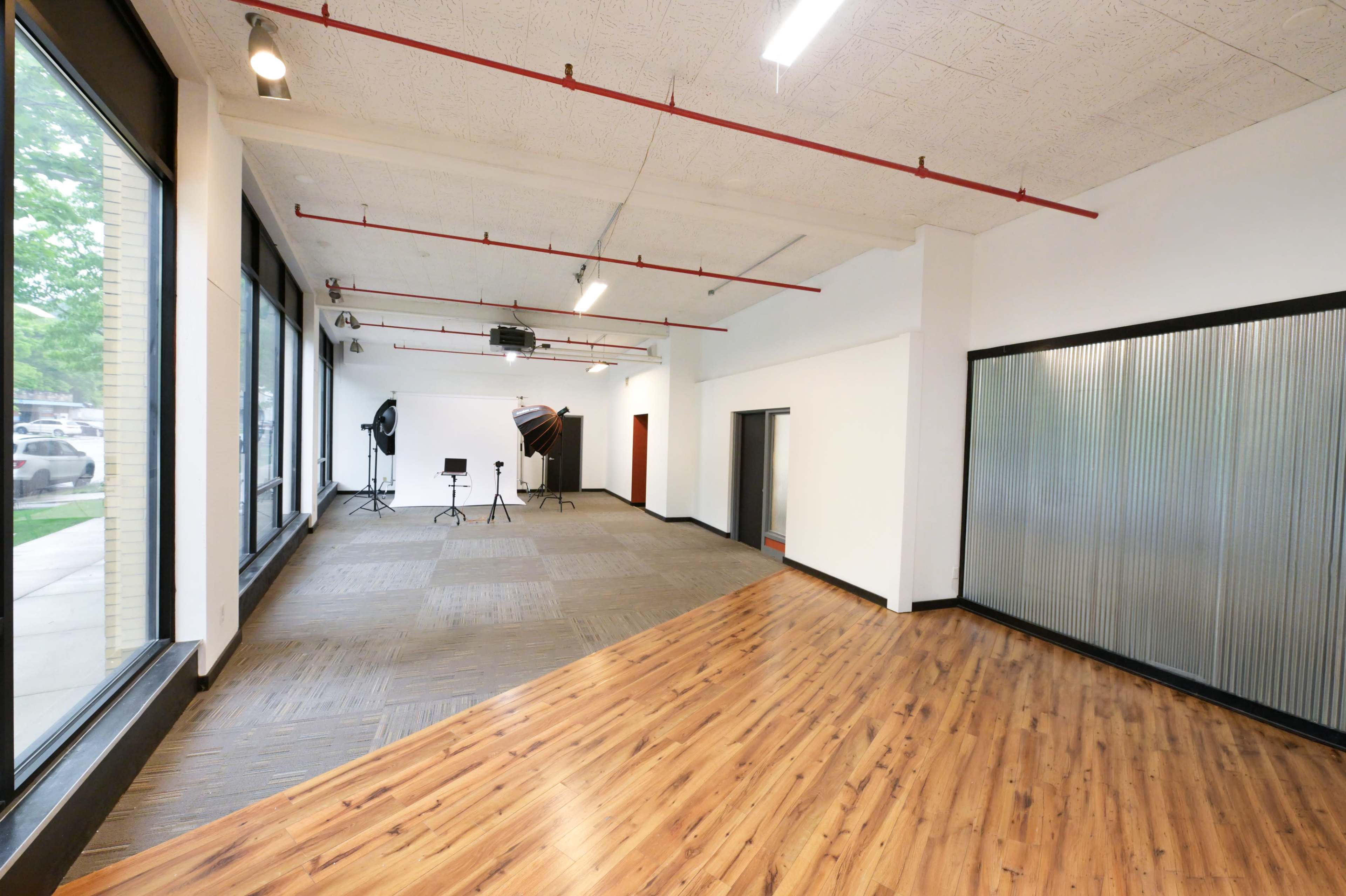 The image shows an empty studio space with wooden flooring, large windows, and equipment stands positioned near the back.