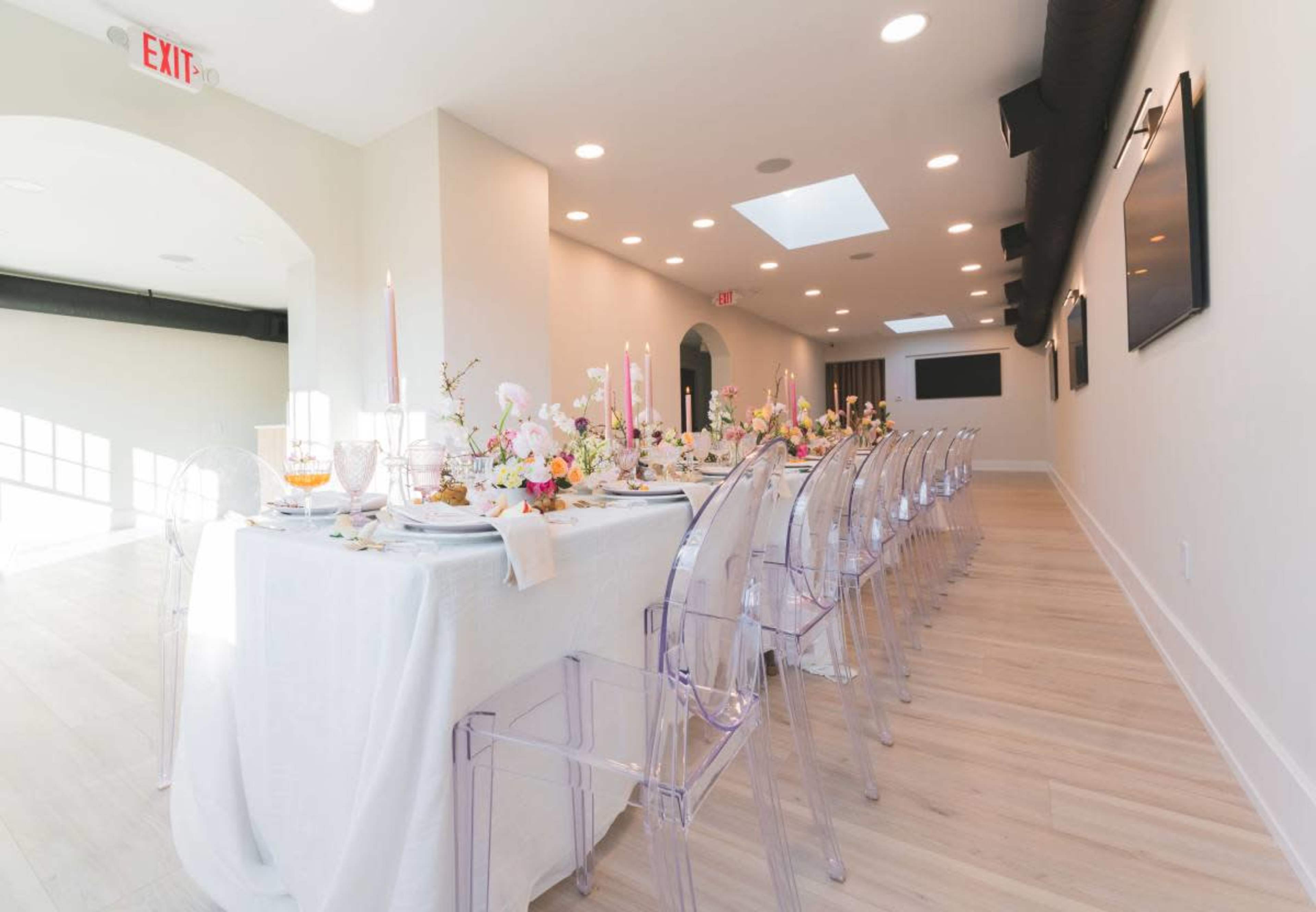 A long dining table is set with pastel decorations and clear acrylic chairs in a well-lit, modern room.