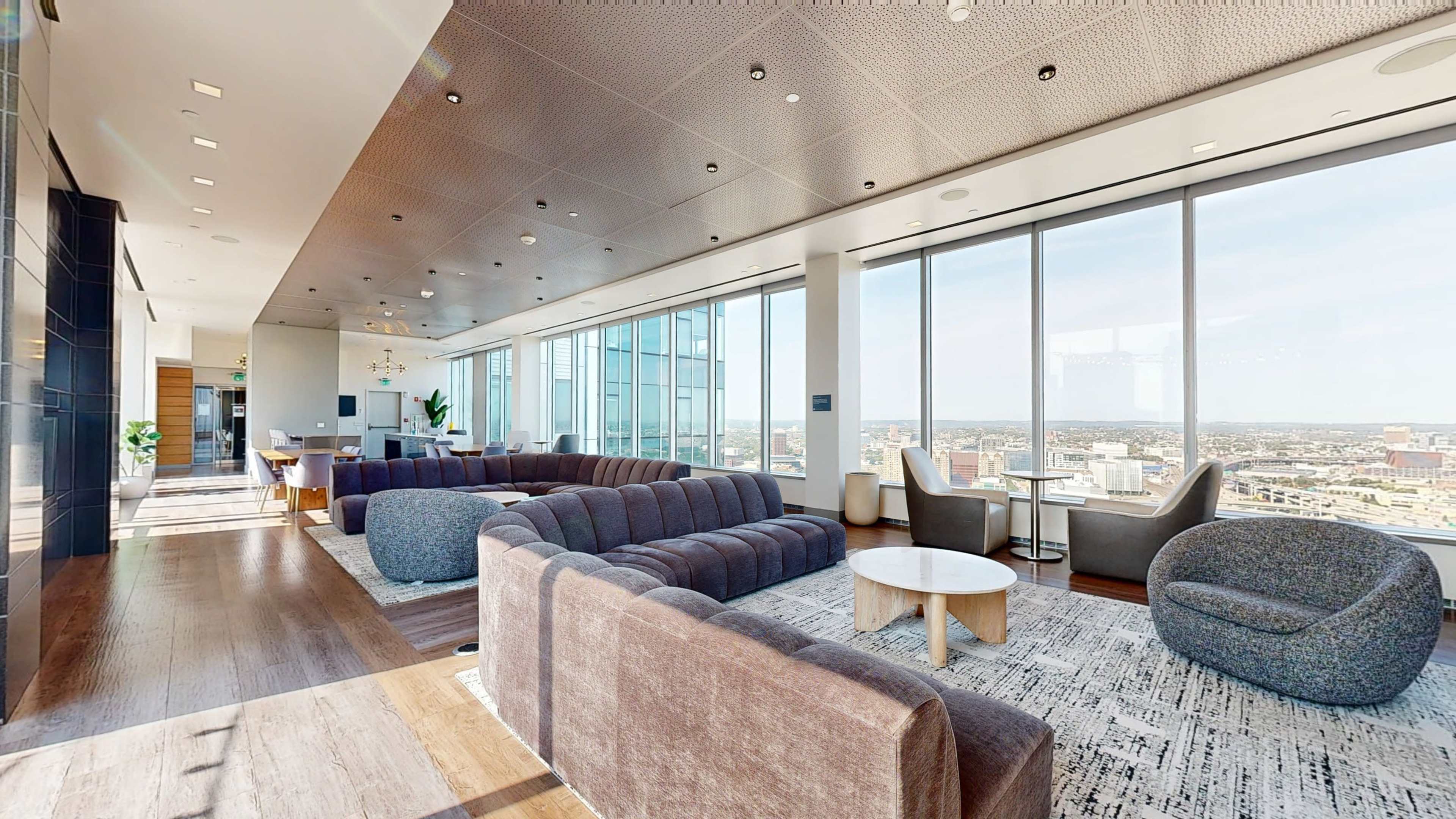 The image shows a modern, spacious lounge area with large windows, a variety of seating options, and a view of the city skyline.
