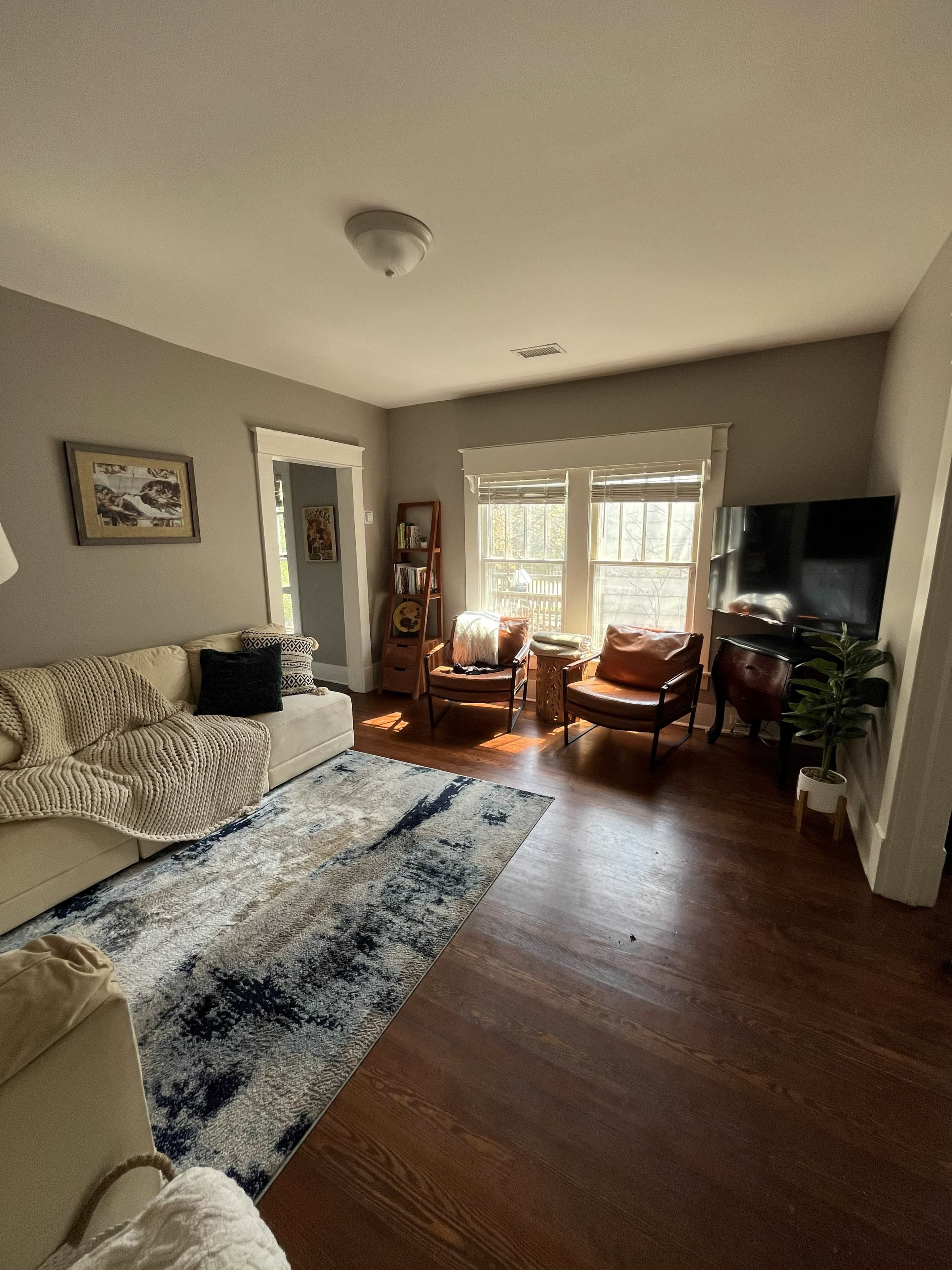 The living room features a light-colored sofa, two leather chairs, a wooden bookshelf, and a television against a wall, with sunlight streaming through the windows.
