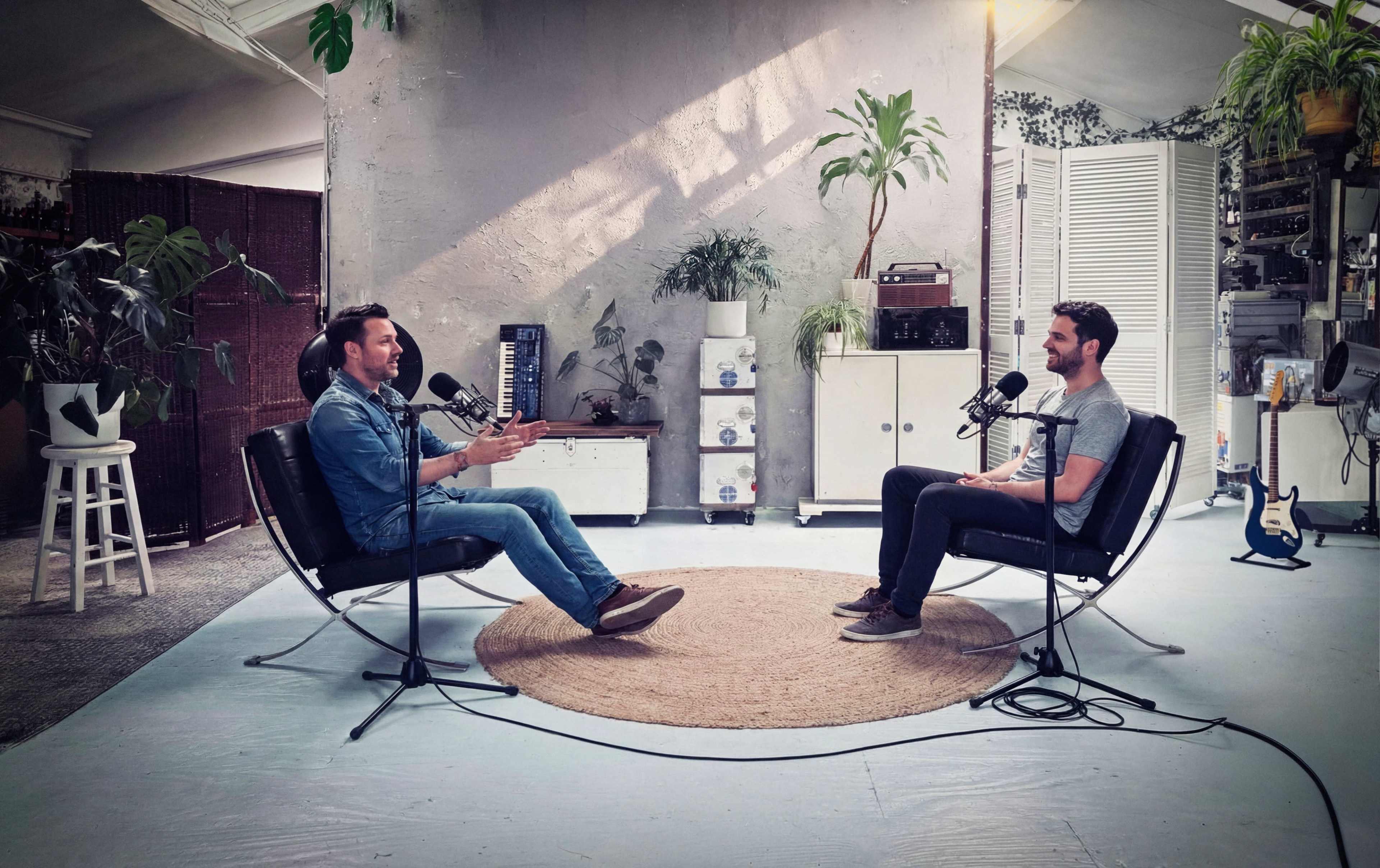 Two men are seated in modern chairs facing each other in a well-lit studio surrounded by plants, with microphones positioned between them.