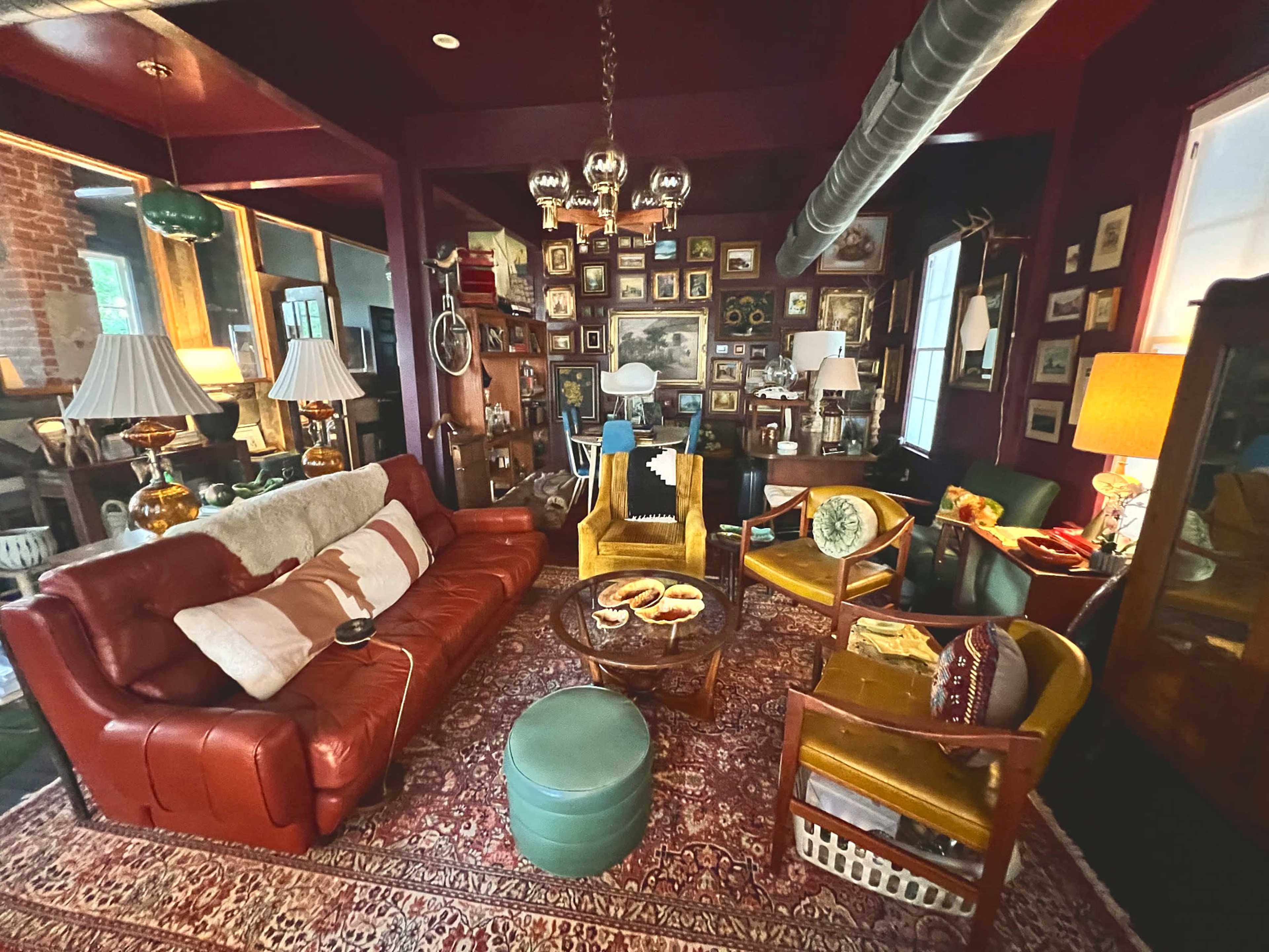 The image shows a cozy living room with a red leather sofa, colorful armchairs, a round coffee table, and walls adorned with various framed pictures.