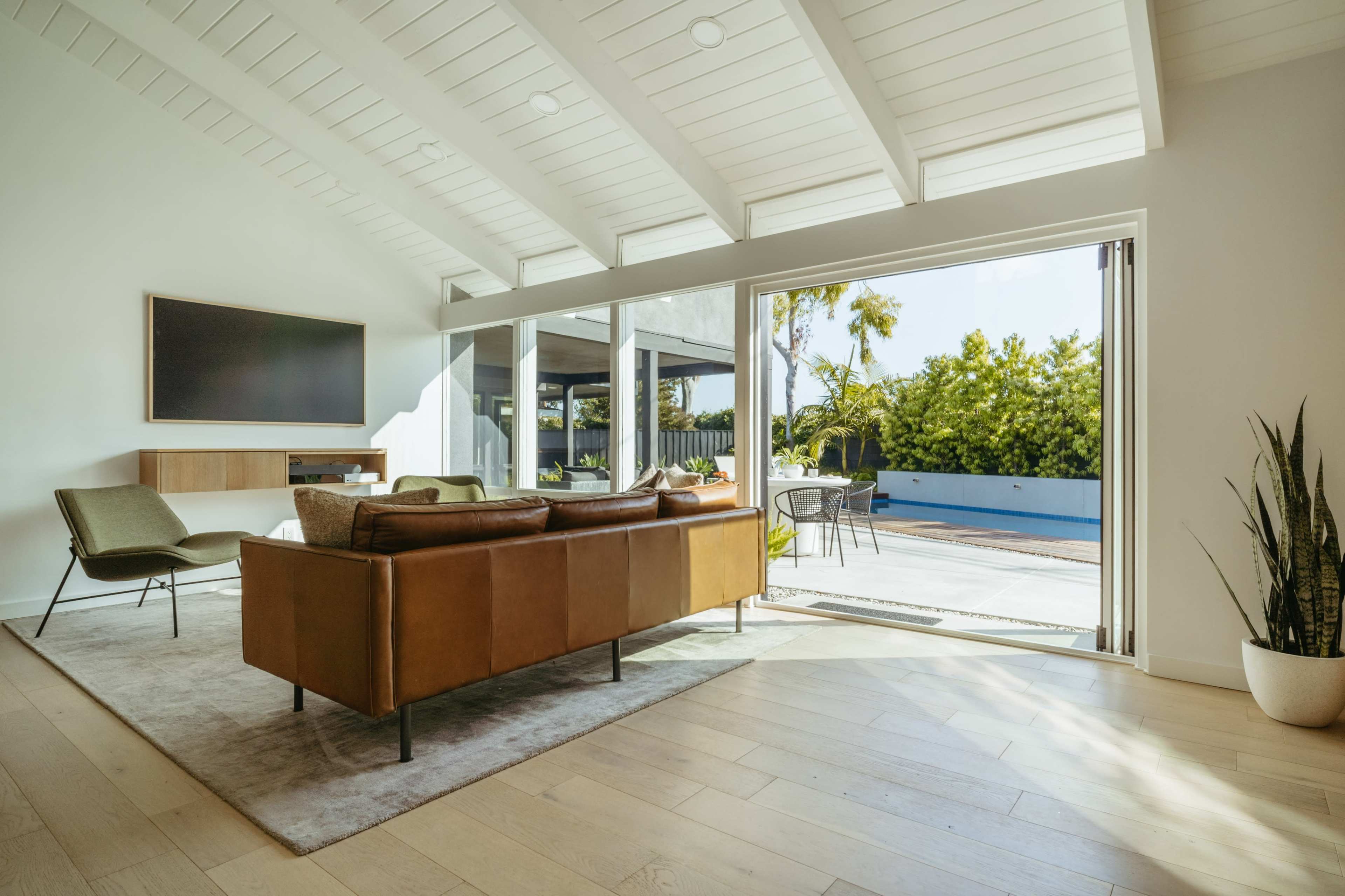 A modern living room features a brown leather sofa, a flat-screen TV on the wall, and large glass doors that open to a patio with outdoor seating and greenery.