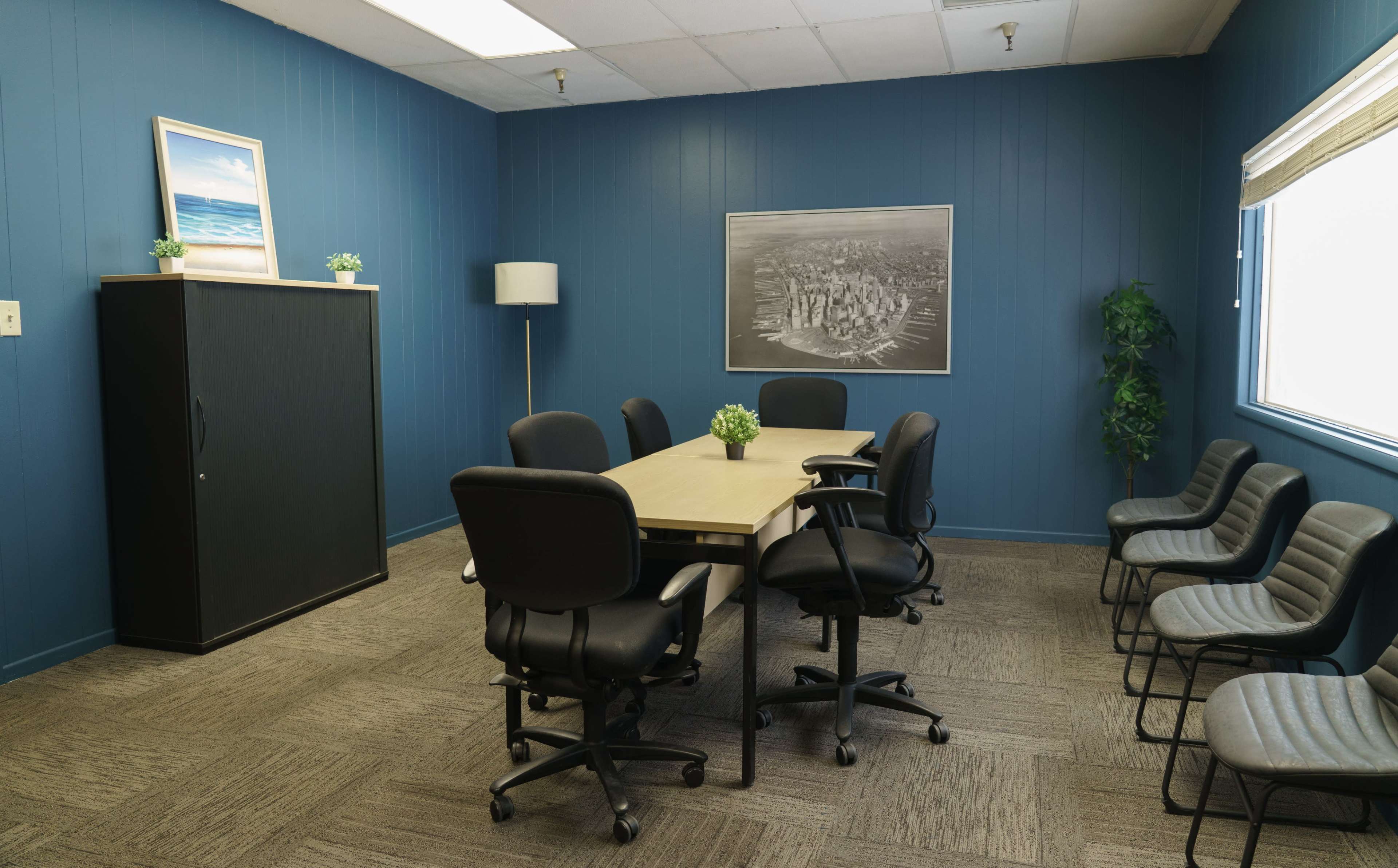 The image shows a conference room with a central table surrounded by rolling chairs, a black cabinet, and two plants along the walls.