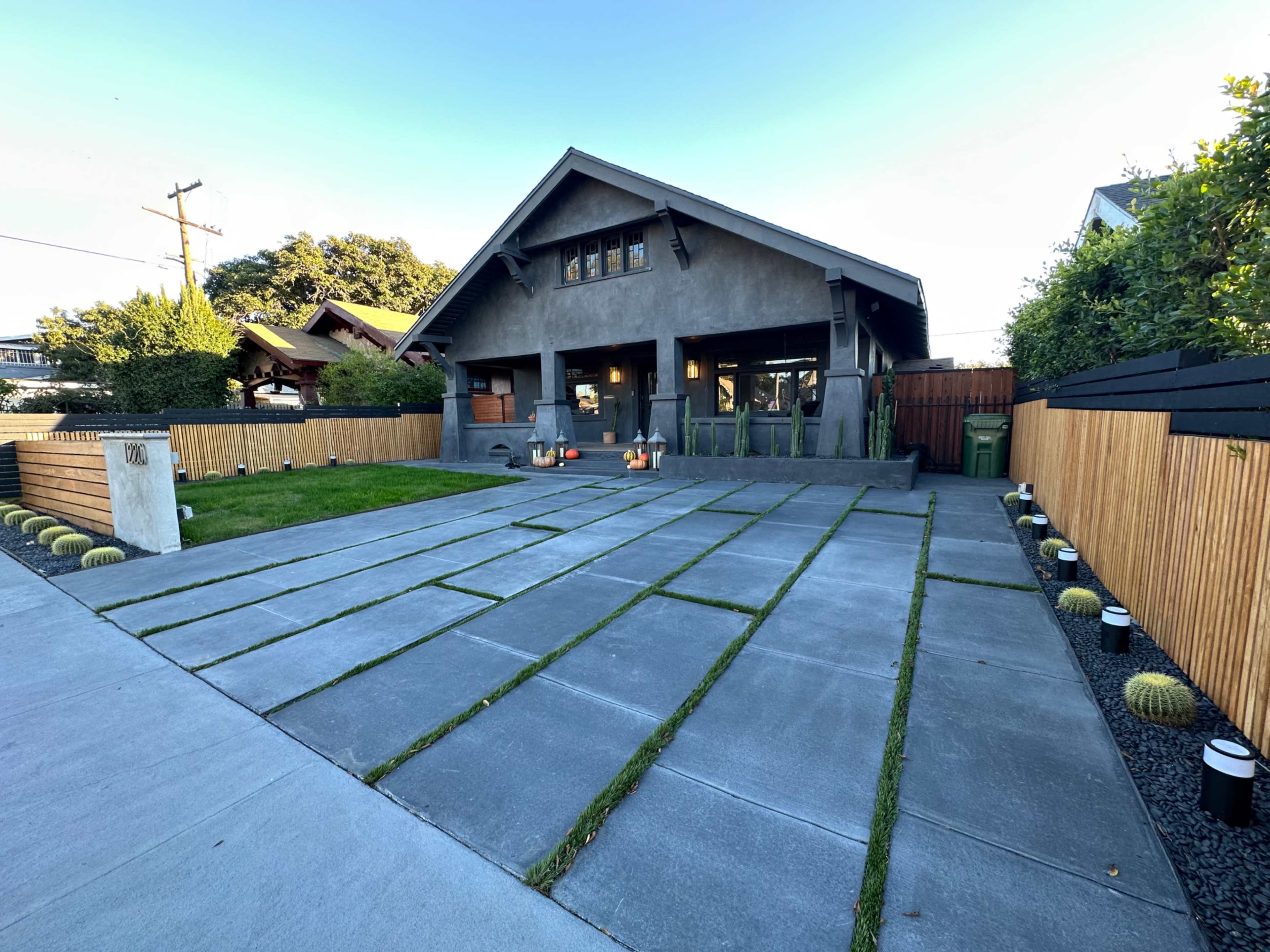 The image shows a modern house with a concrete driveway featuring grassy strips and landscaped areas, surrounded by a wooden fence and various plants.