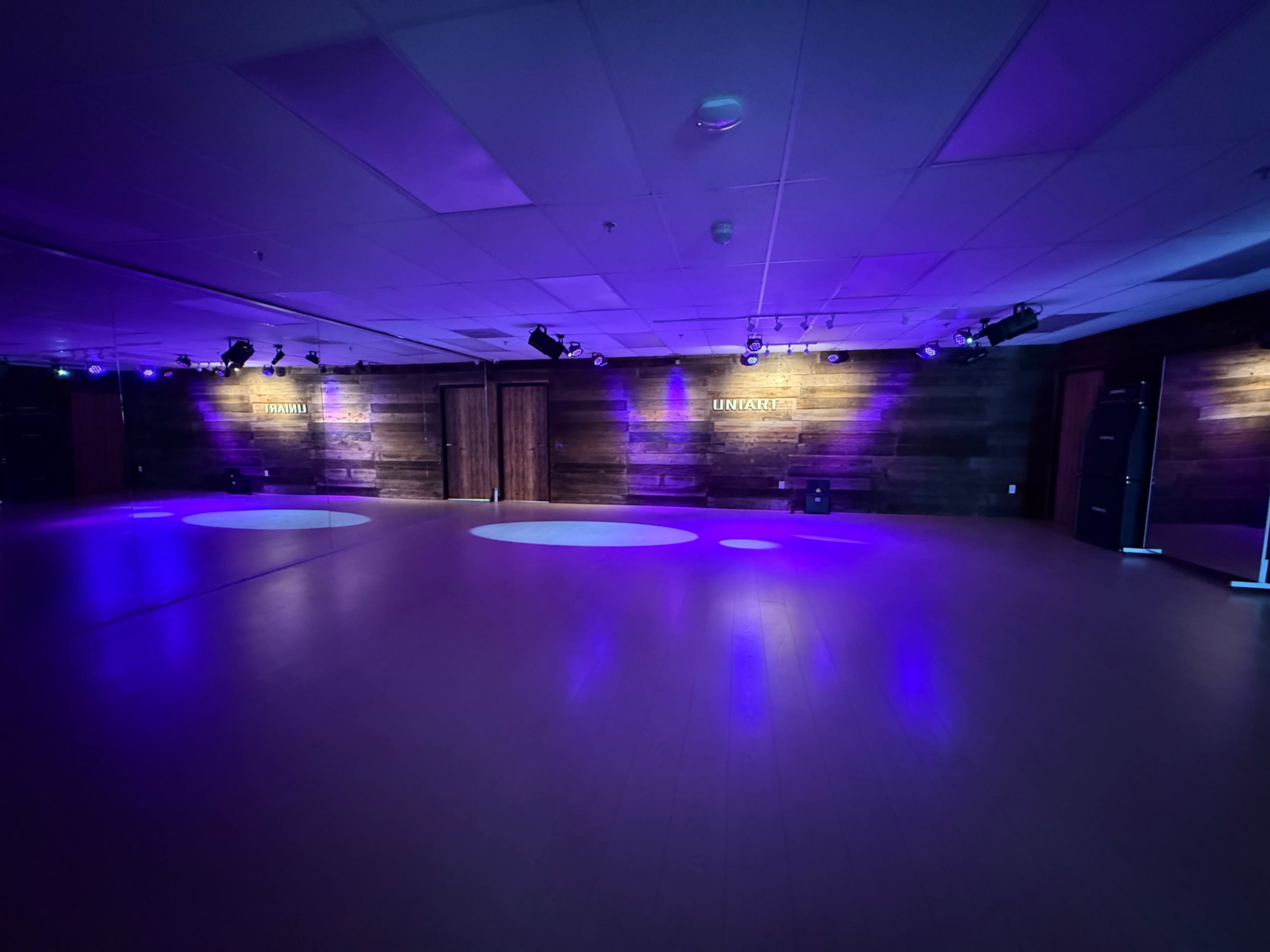 The image shows an empty dance studio with purple lighting and wooden walls, featuring circular light spots on the floor.