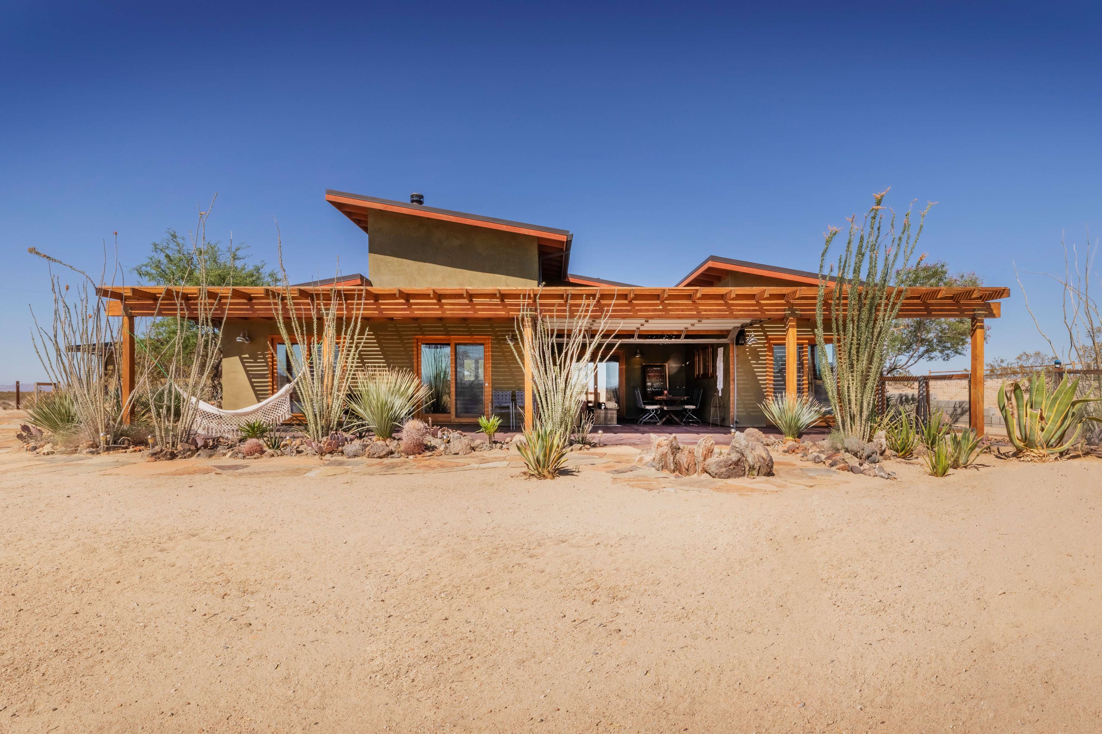 A modern desert house with wooden accents and large windows is surrounded by cacti and gravel landscaping.