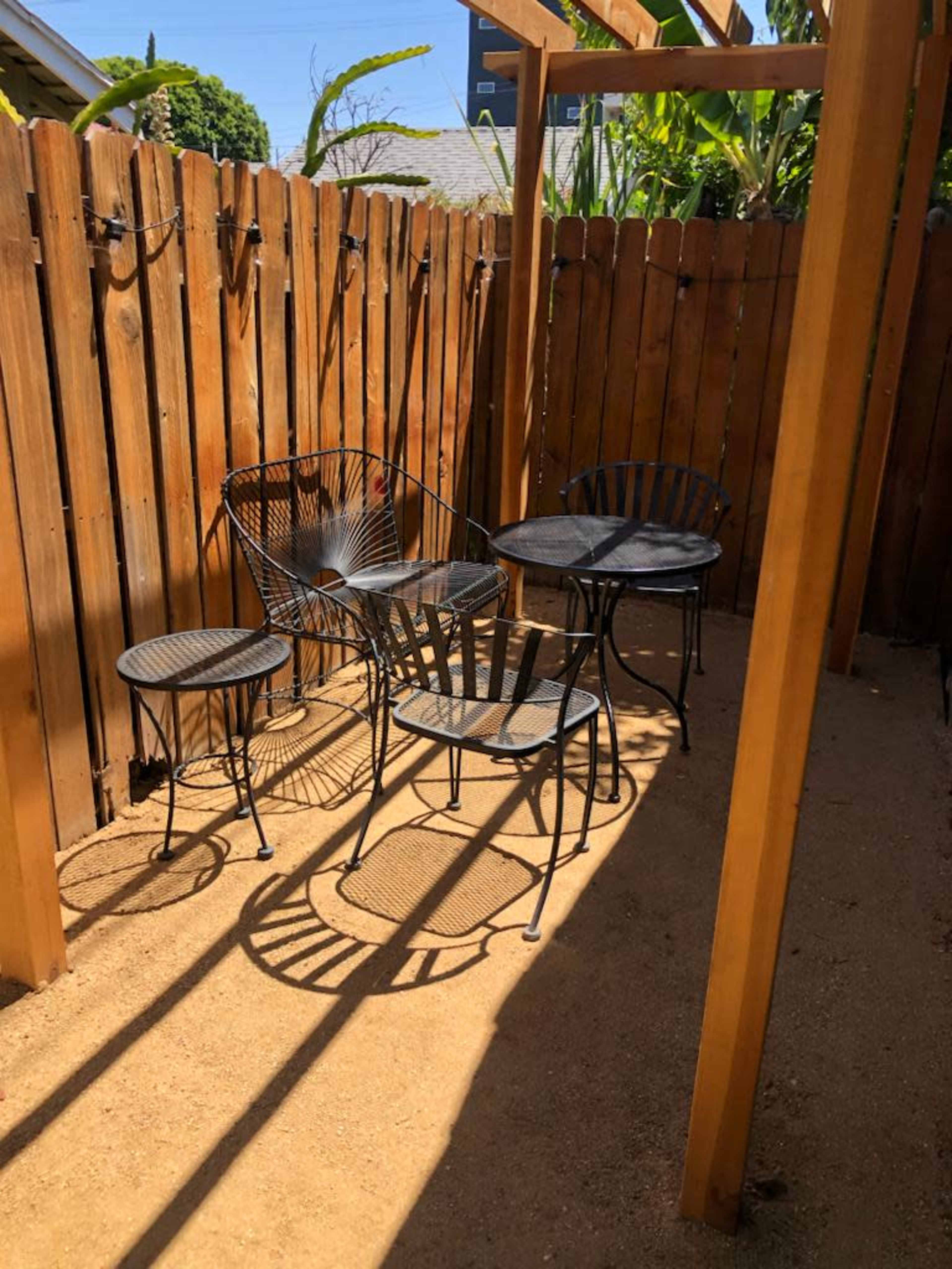 A small outdoor seating area features two black metal chairs, a round table, and a small side table on sandy ground, enclosed by a wooden fence and shaded by a canopy.
