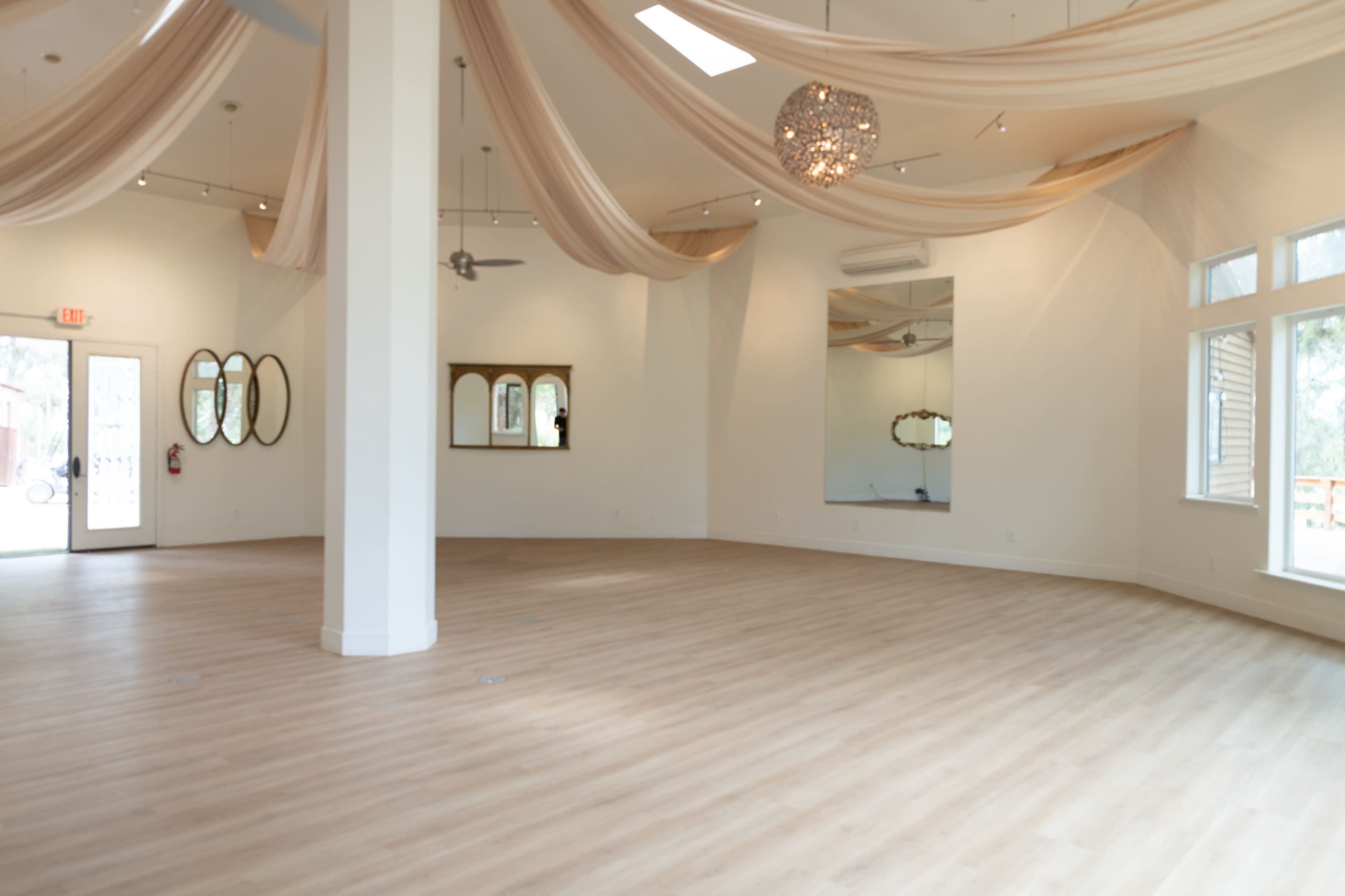 The image shows a spacious, well-lit room with light wood flooring, elegant draped fabric on the ceiling, and decorative mirrors on the walls.