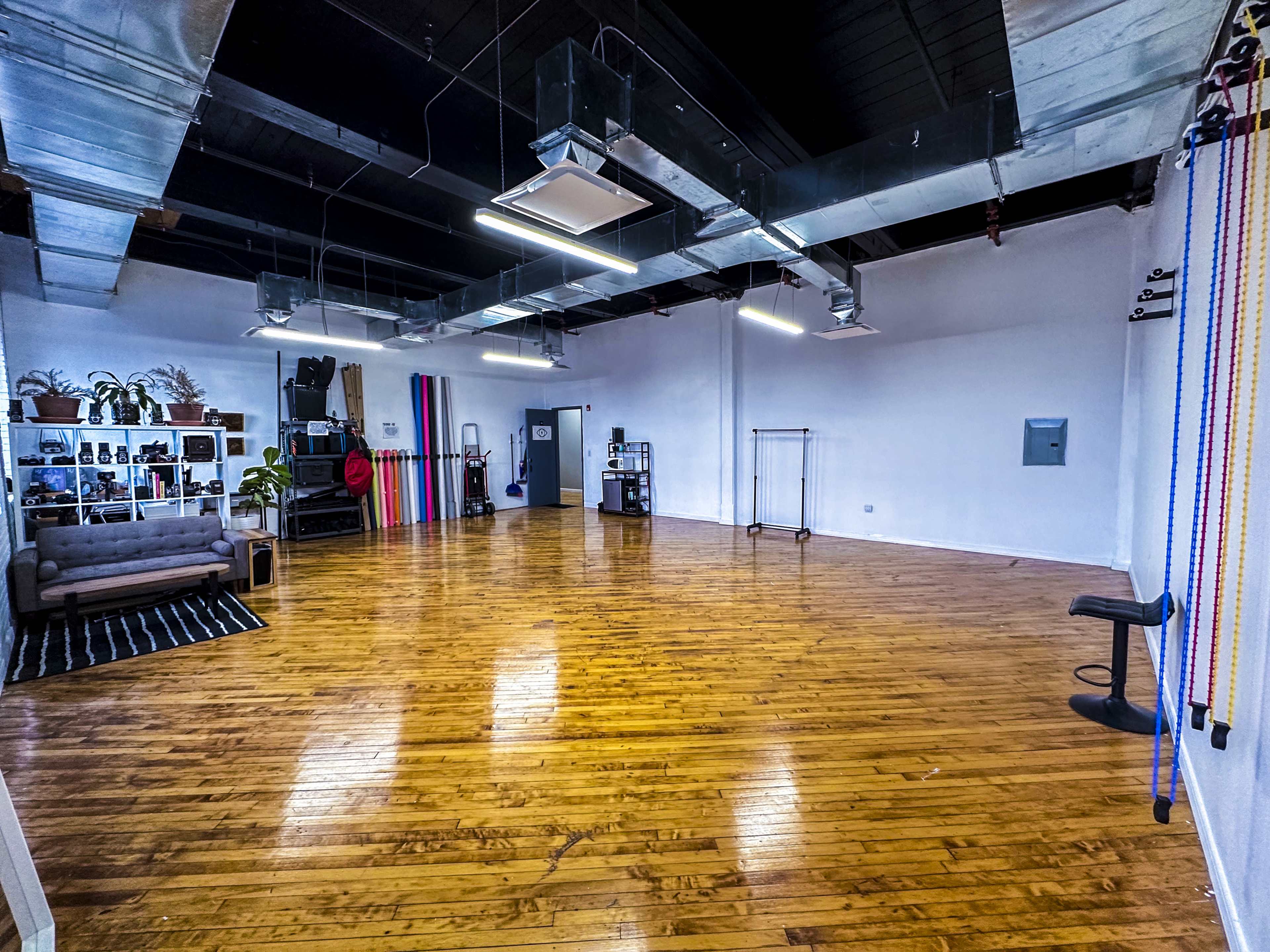 An empty studio with wooden floors, large windows, and minimal furniture, including a couch and a clothing rack.