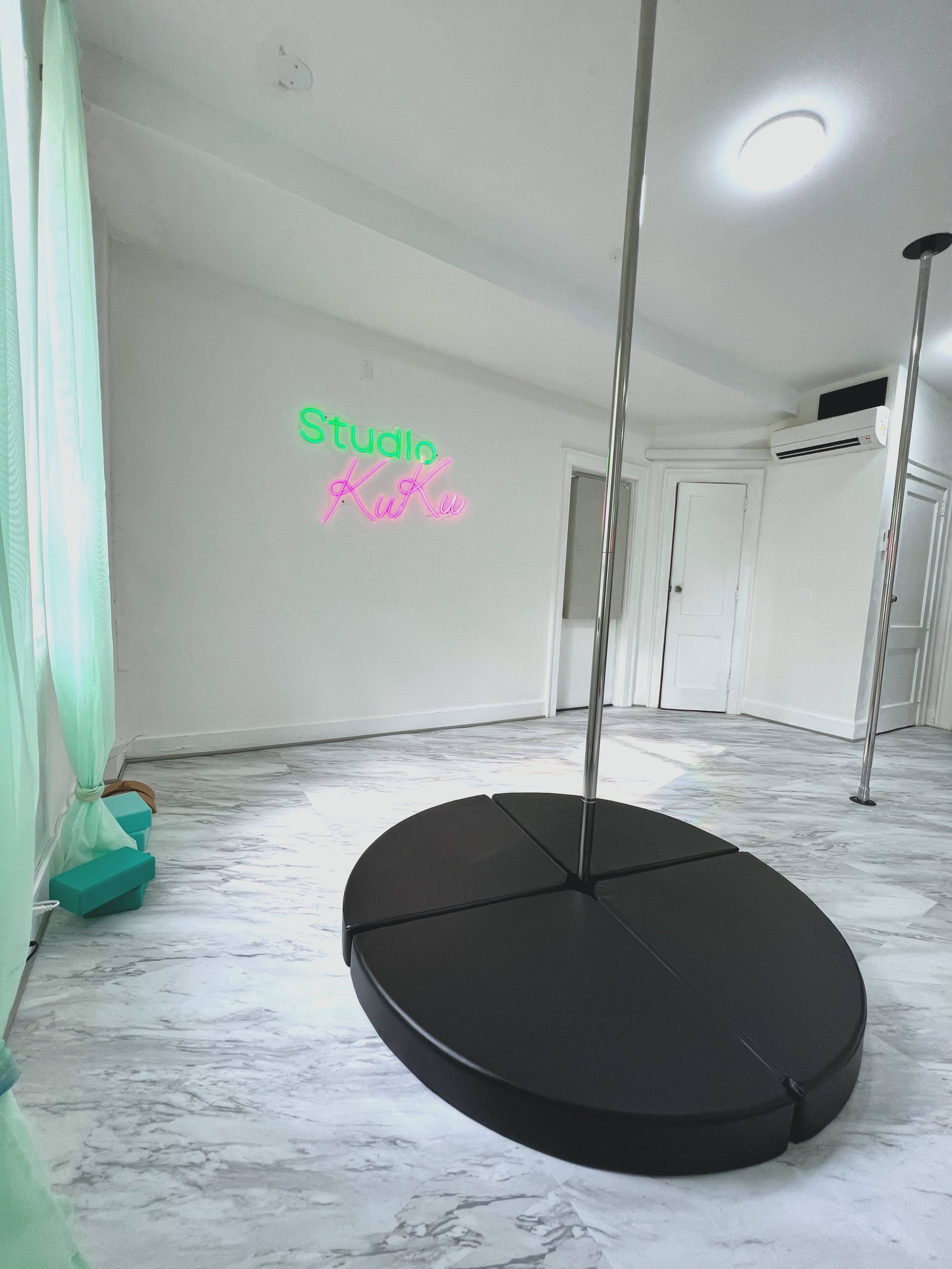 A spacious studio with a black pole on a large floor mat and a neon sign that reads "Studio Kiki" on the wall.