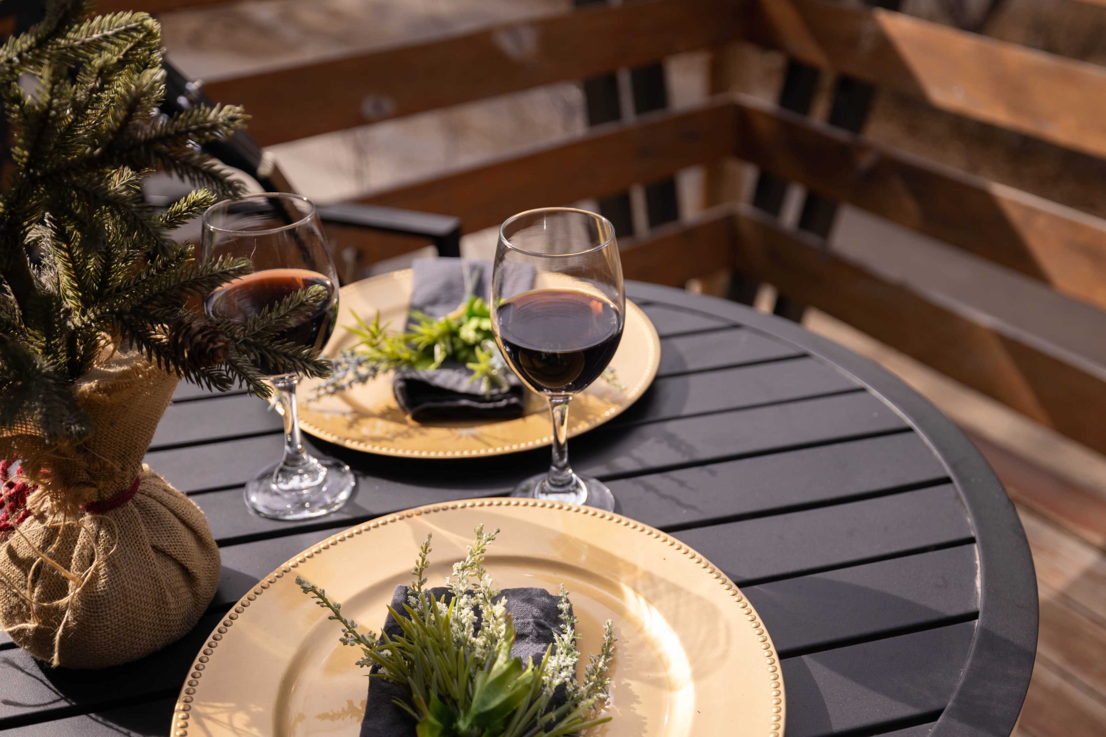 A table set for two with wine glasses and plates adorned with greenery sits on a wooden balcony.