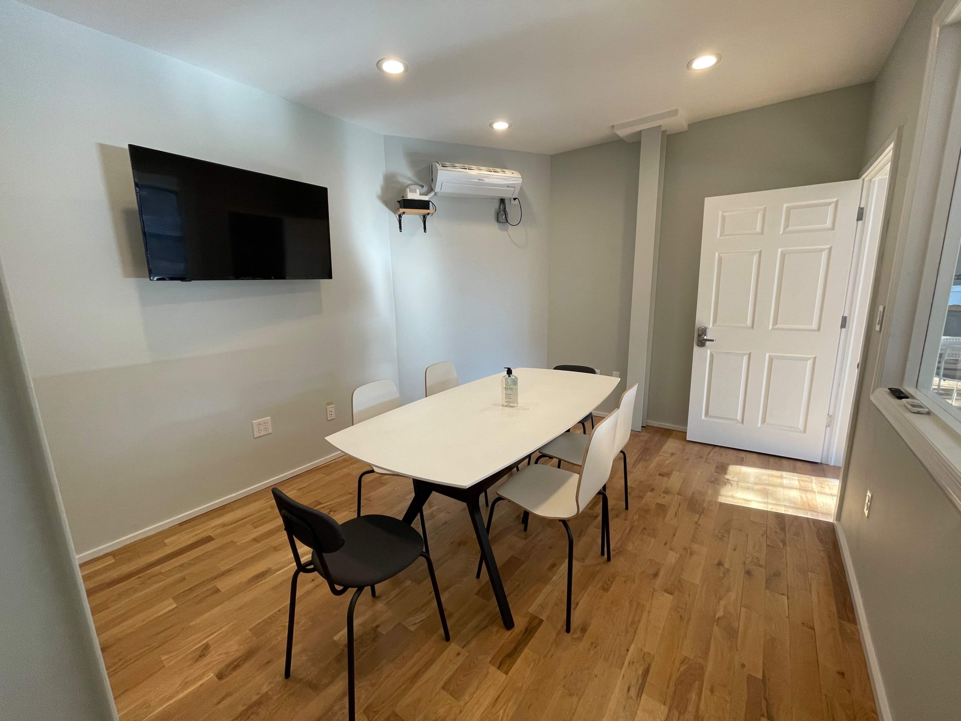 A small meeting room features a rectangular table surrounded by four chairs, a wall-mounted television, and a door leading to another space, with wood flooring and light-colored walls.