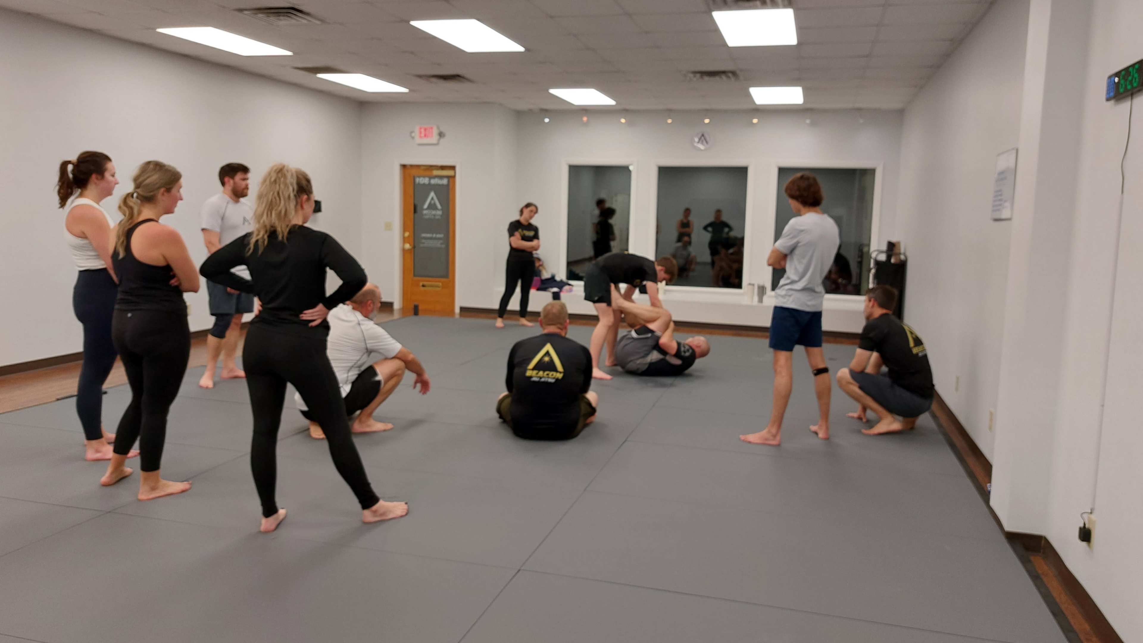 A group of individuals is participating in a martial arts training session on a gray mat inside a bright, mirrored room.