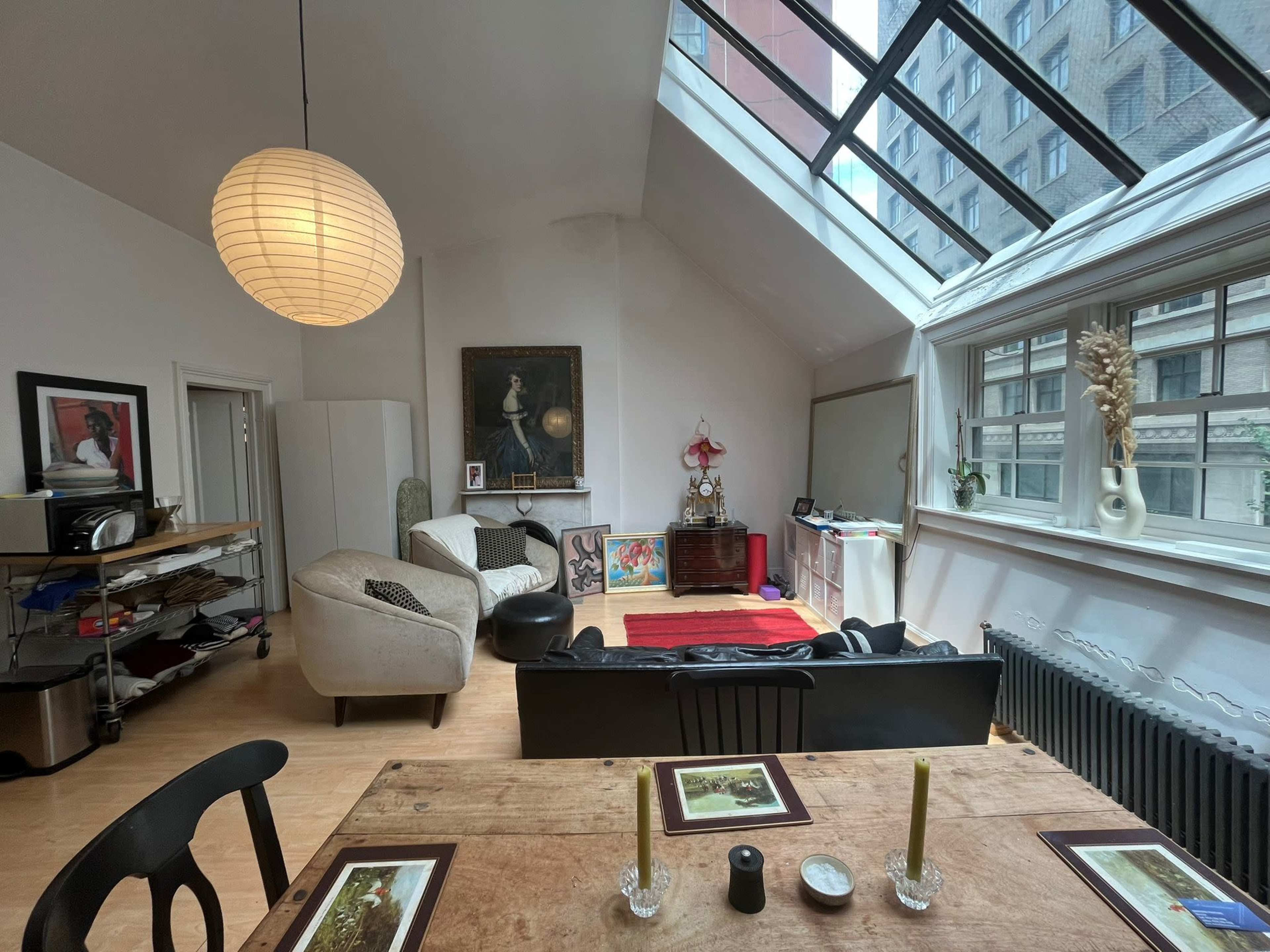 The image shows a modern living room with a skylight, featuring a mix of seating arrangements, artwork on the walls, and a wooden dining table.