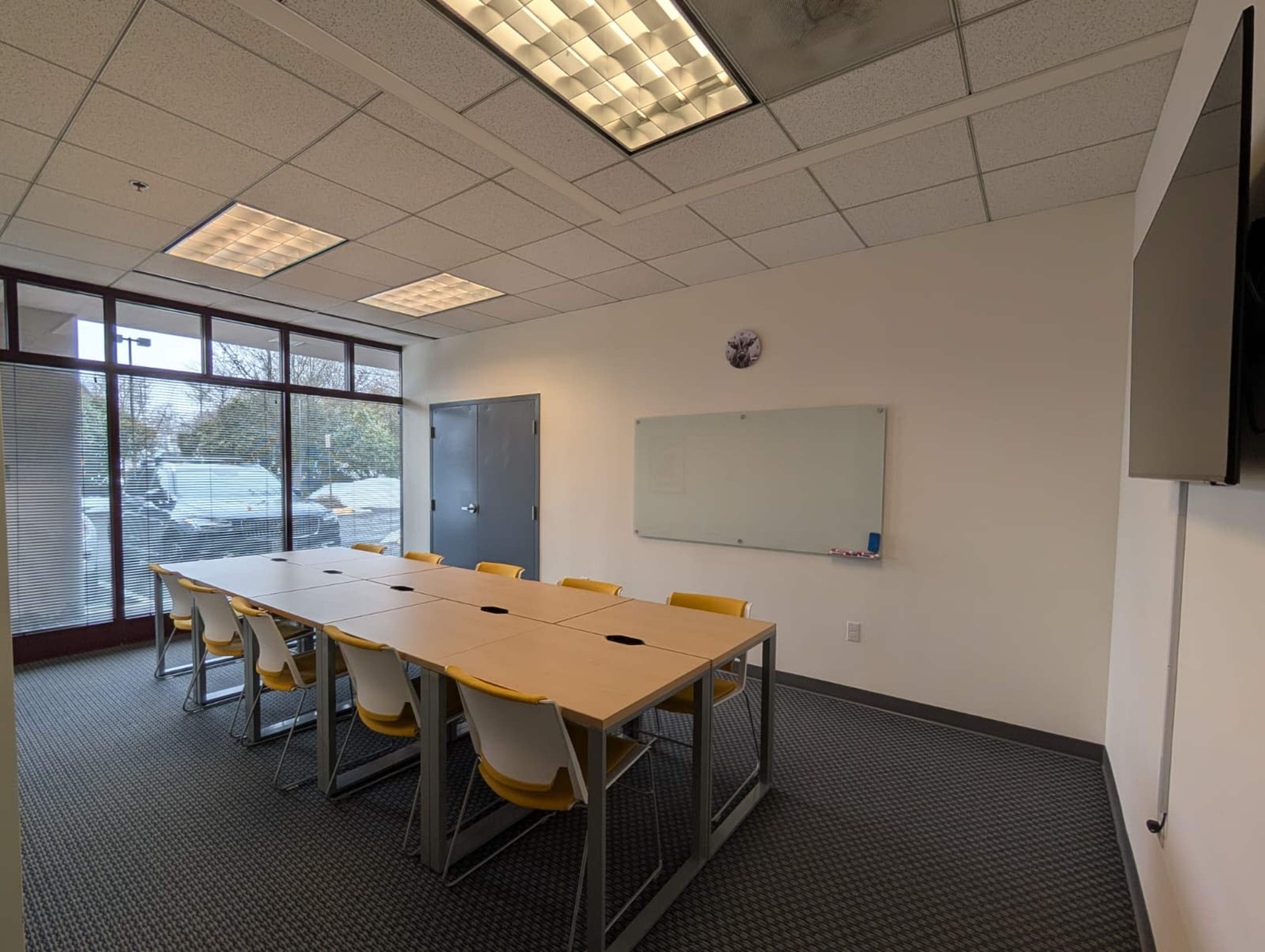The image shows a conference room with a large table surrounded by yellow chairs, a whiteboard, and a wall-mounted TV.