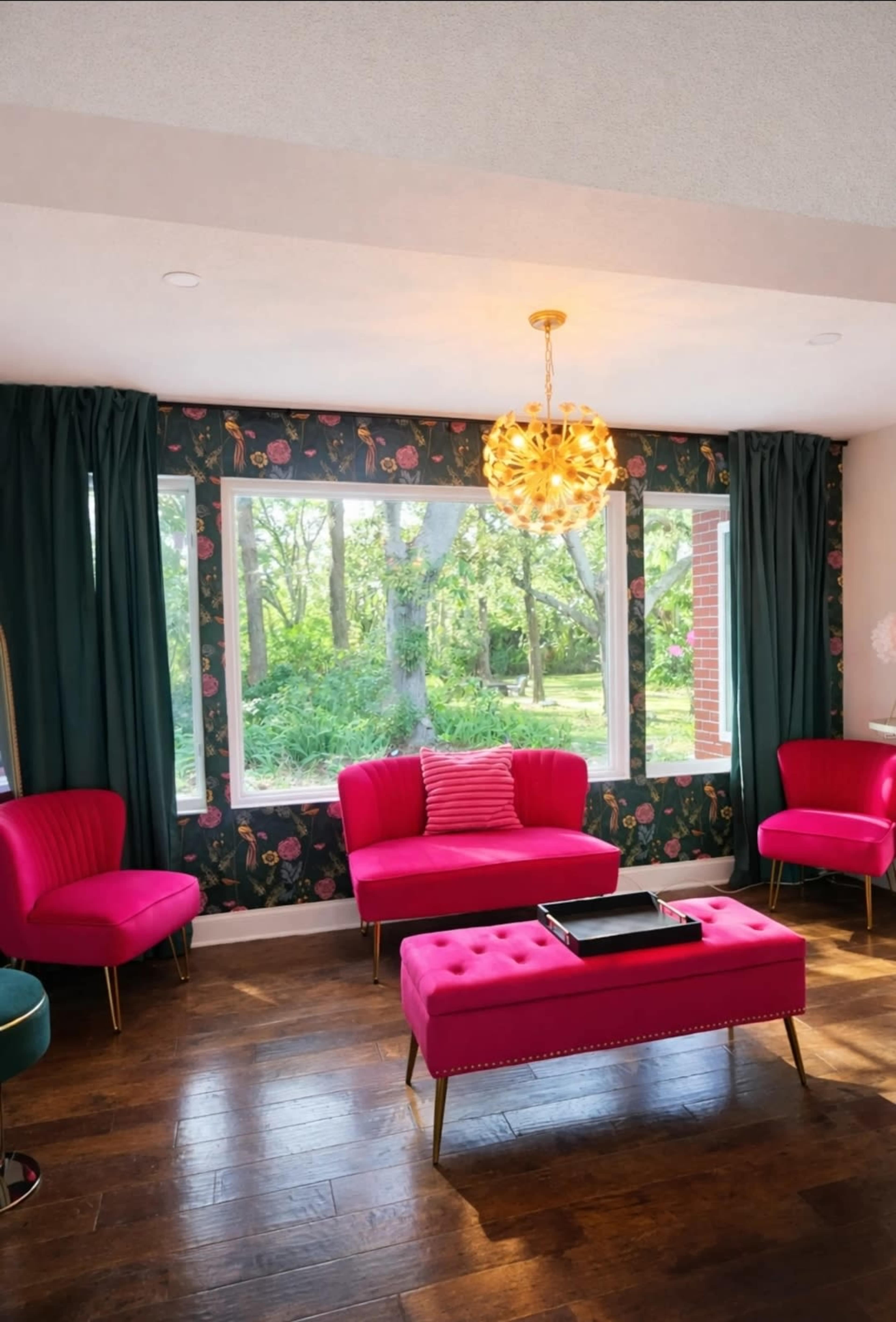 A brightly colored living room features pink chairs and a tufted pink coffee table, with large windows overlooking a green landscape.