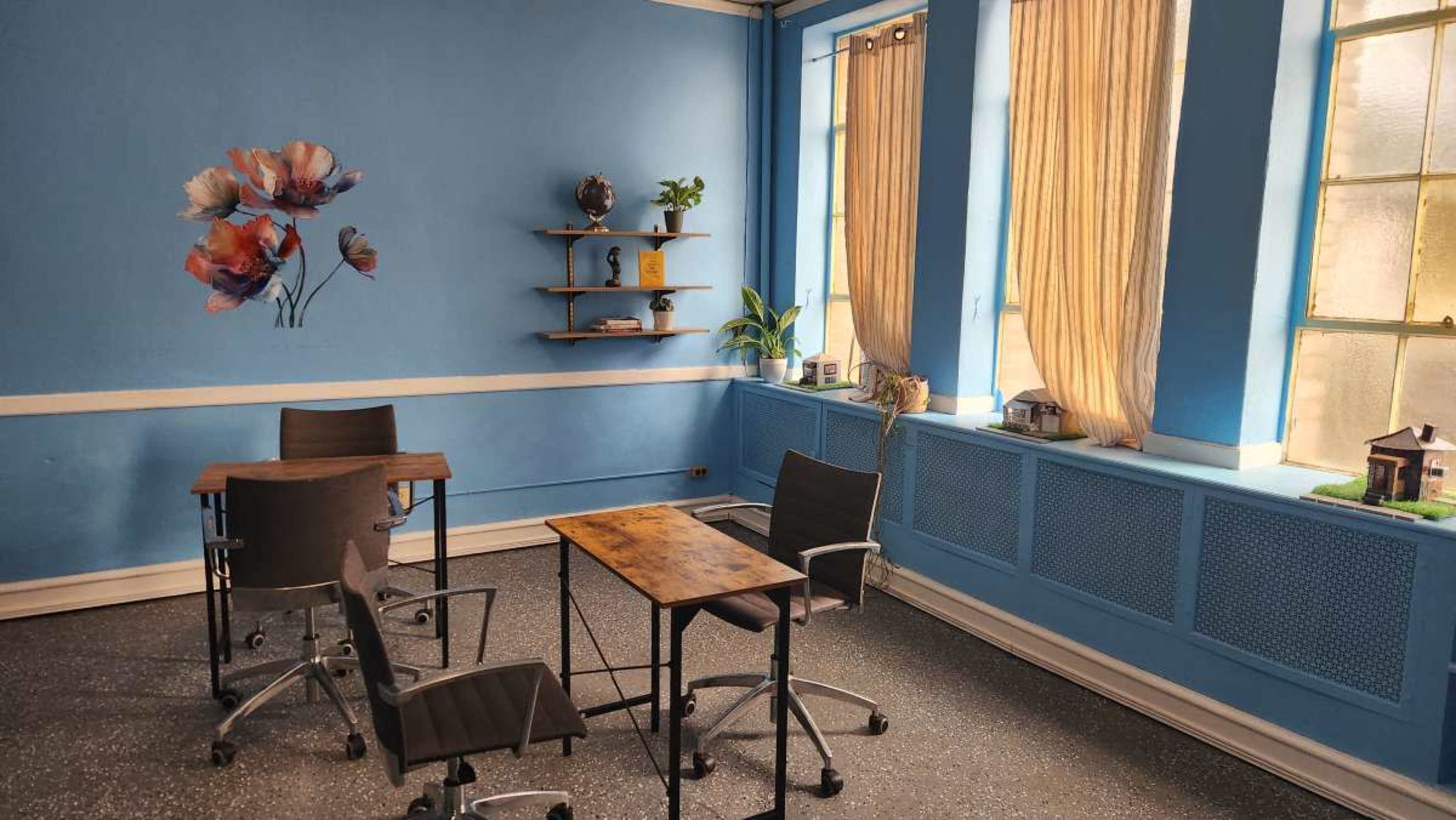 The image shows a bright blue room with two windows, a floral wall design, and furniture consisting of two wooden tables, four chairs, and a shelf with decorative items.